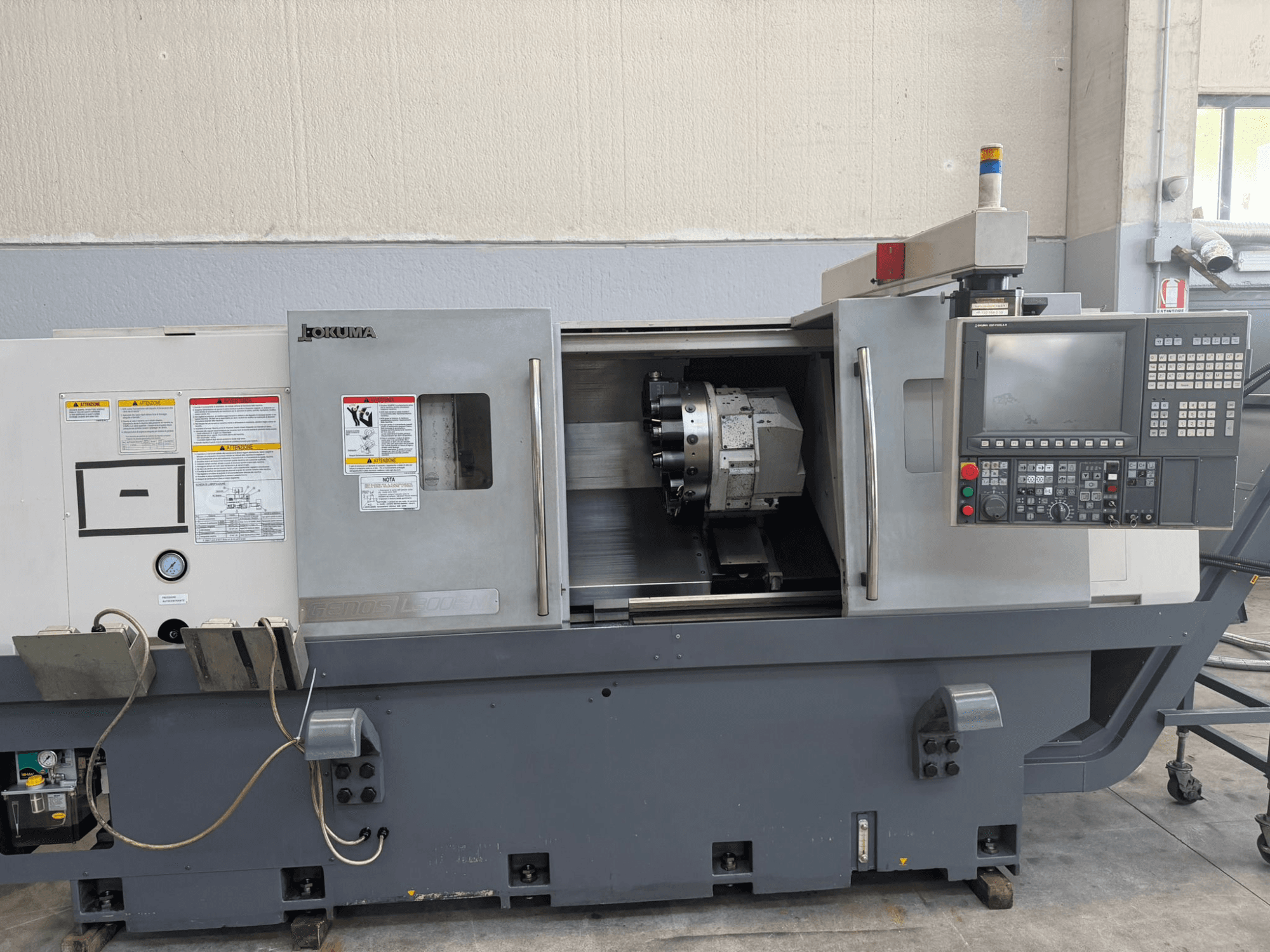 Tornio CNC Okuma, vista frontale, con pannello di controllo, portautensili e avvertenze di sicurezza sul lato.