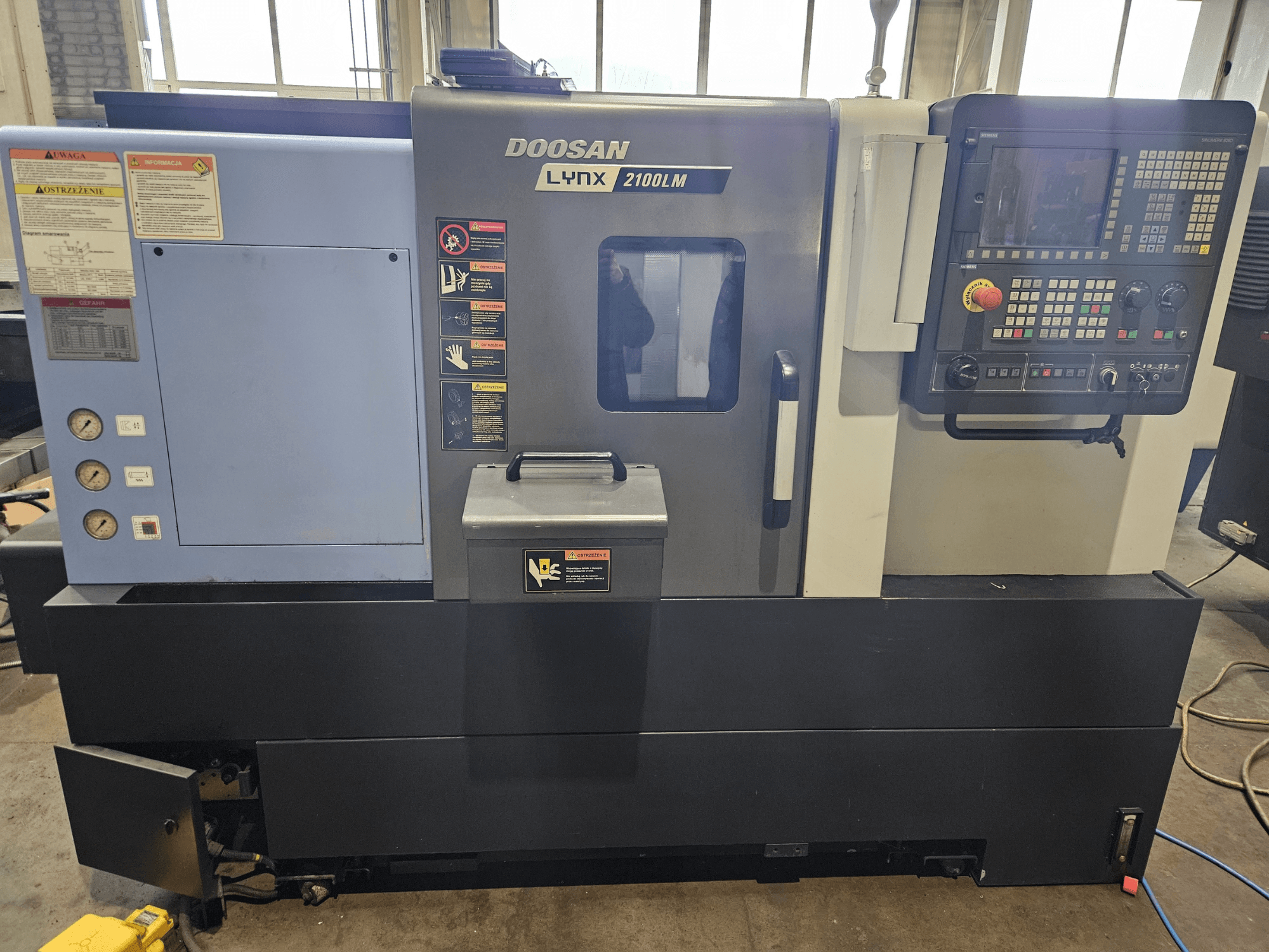 Tornio CNC Doosan Lynx 2100LM, vista frontale, con pannello di controllo, manometri dell'aria ed etichette di sicurezza visibili.