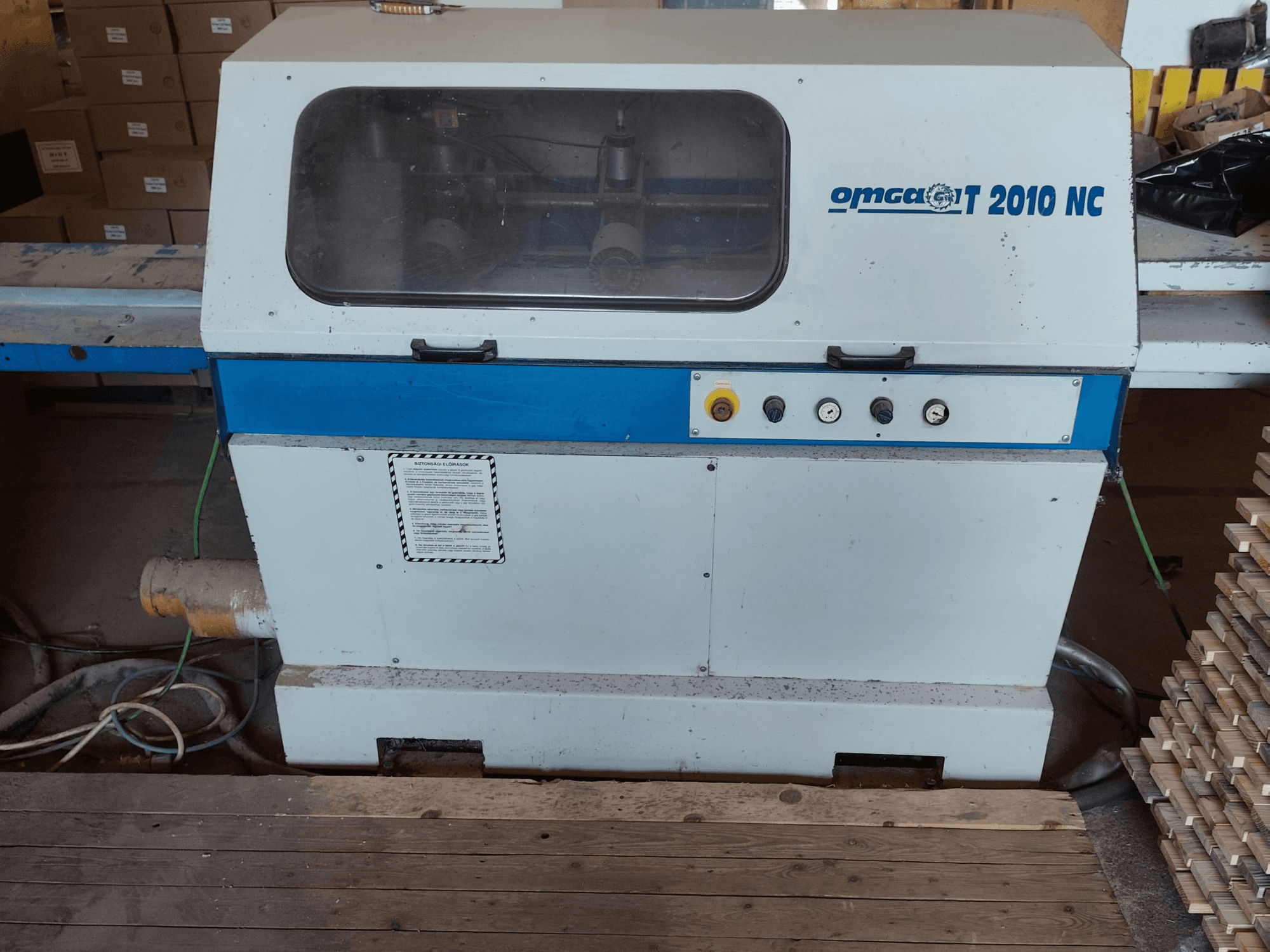 Ottimizzazione della sega trasversale OMGA T 2010 NC, vista frontale, con pannello di controllo e copertura trasparente, con legname accatastato accanto.