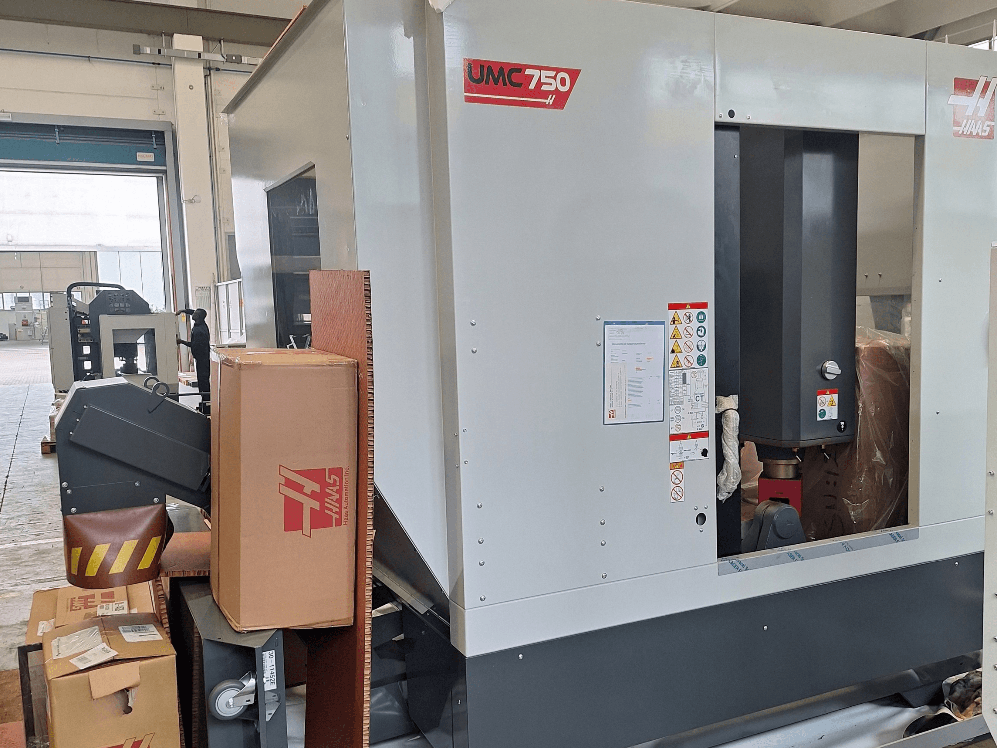 Centro di lavorazione universale HAAS UMC-750, vista laterale, che mostra il pannello di controllo e il vano porta-utensili con etichette di avvertenza e imballaggio.