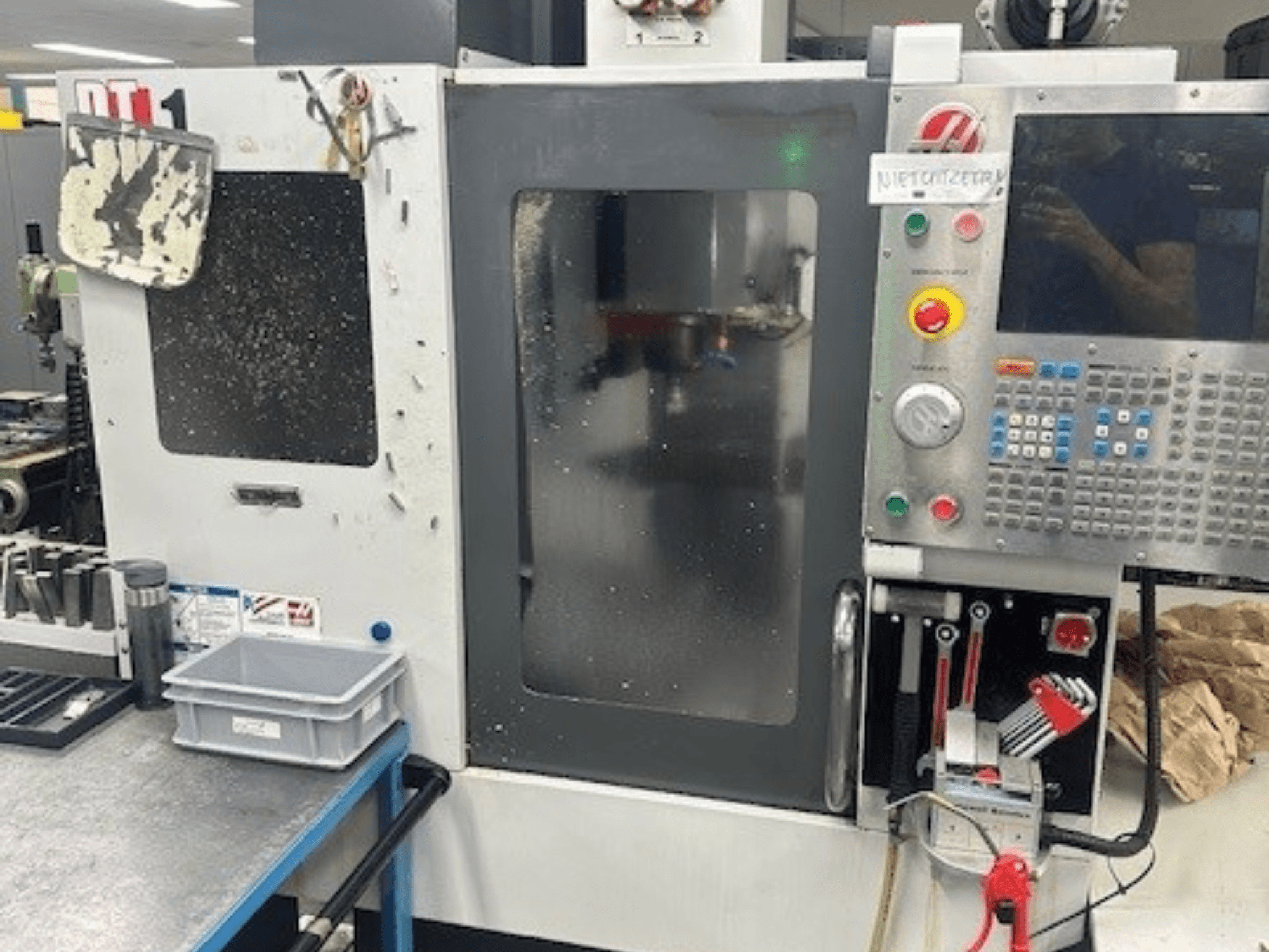 Centro di lavoro verticale Haas DT-1, vista frontale con pannello di controllo, sporco visibile e vassoio portautensili sul banco di lavoro.