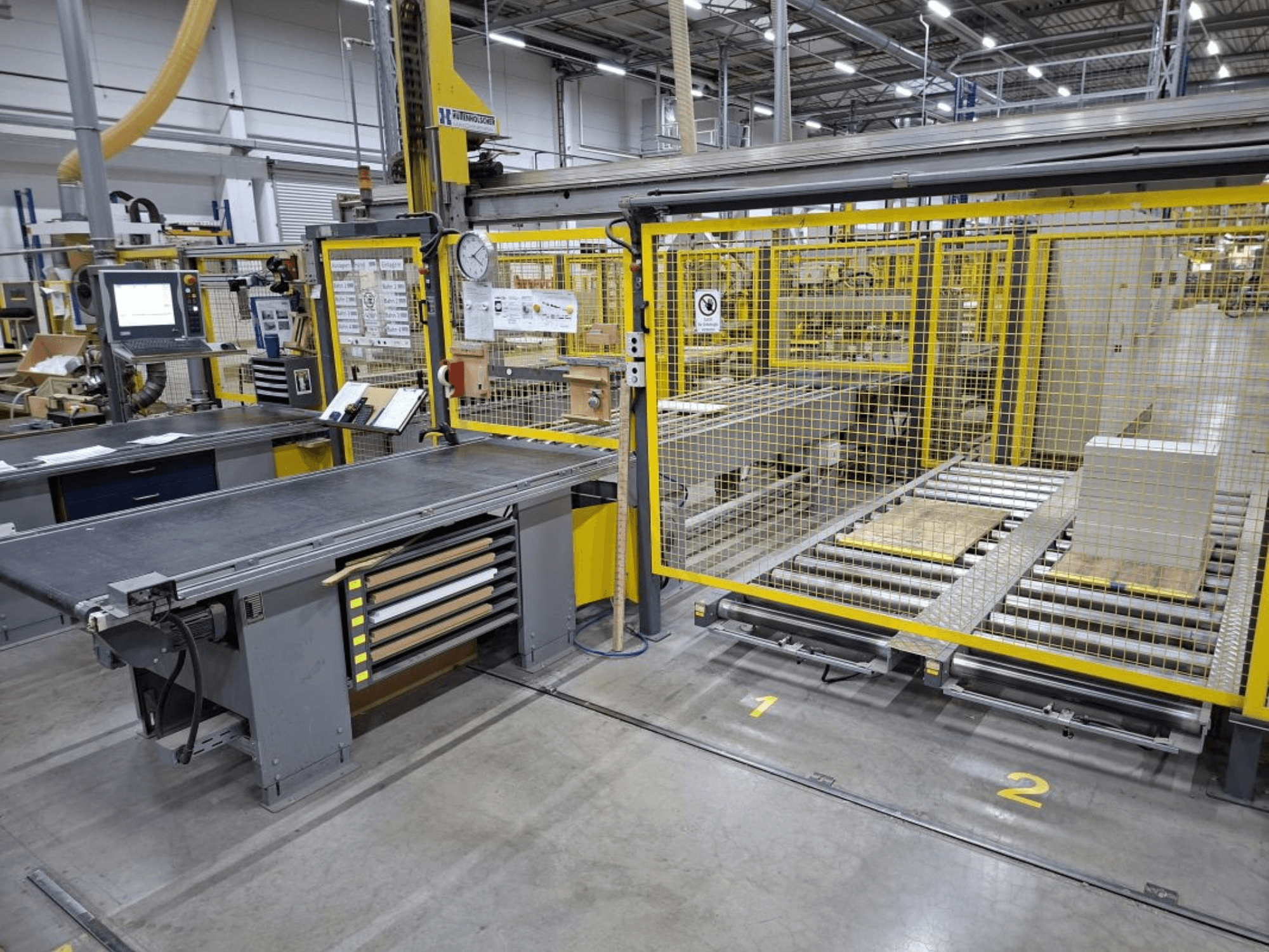 Area di lavoro industriale con sistema di trasporto e pannello di controllo; vista laterale con macchinari e recinzione di sicurezza.