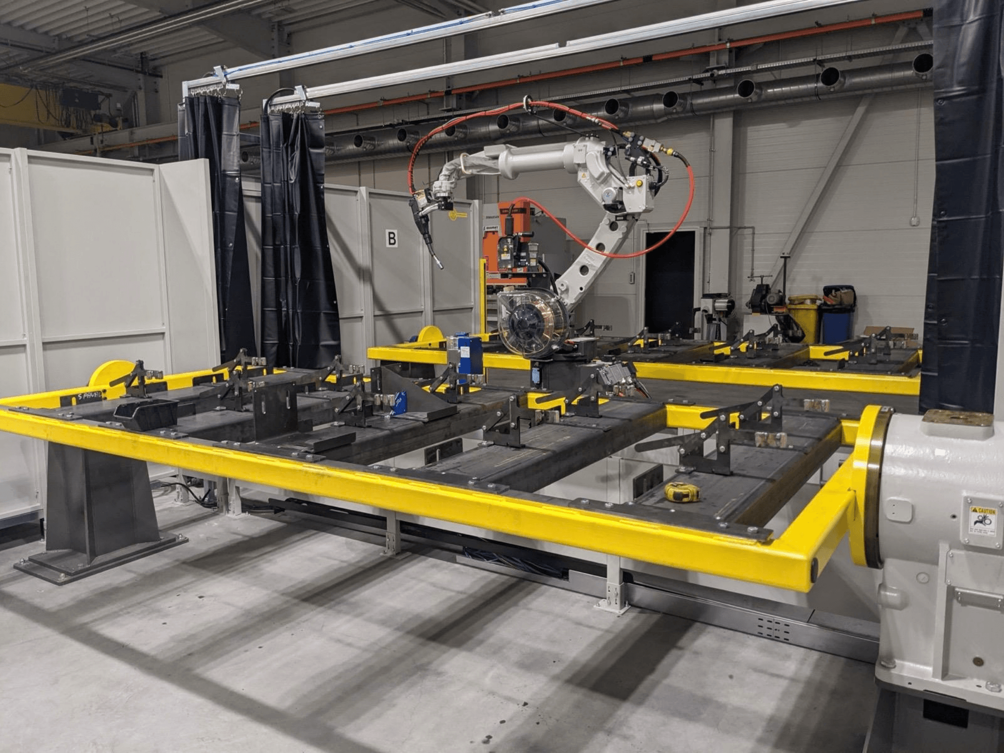 Vista frontale del braccio robotico PANASONIC TAWERS TM-2000WG3 posizionato sopra un tavolo da lavoro giallo su cui sono disposte parti metalliche da assemblare.
