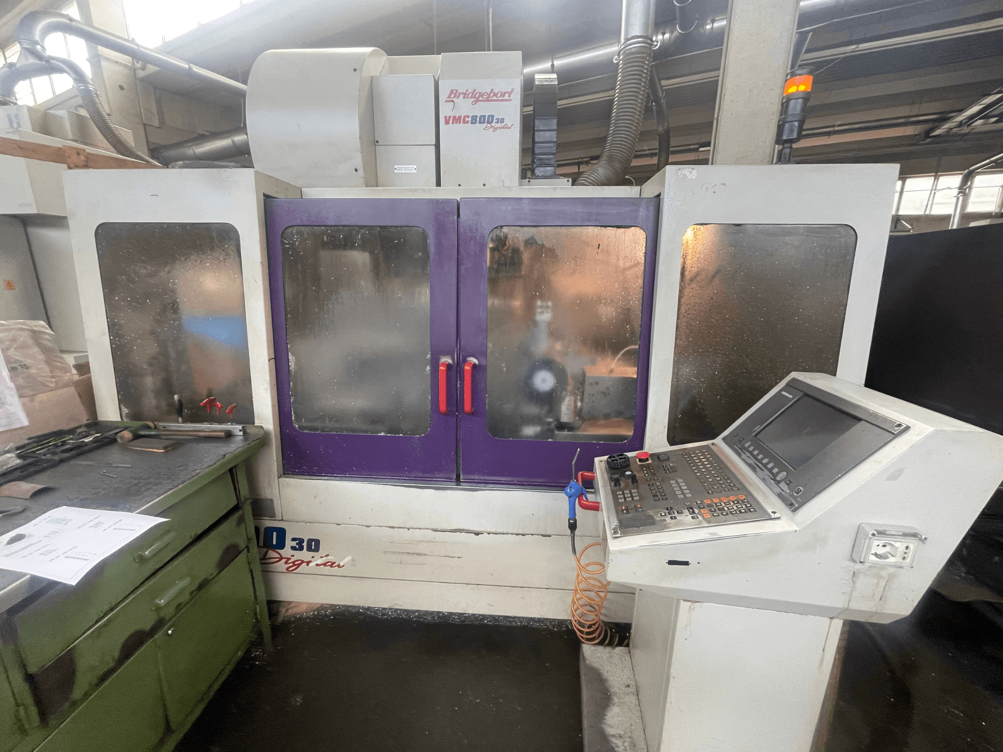 Centro di lavorazione verticale Bridgeport VMC 800/30, vista frontale, con pannello di controllo e sportelli viola in primo piano; utensili sulla superficie adiacente.