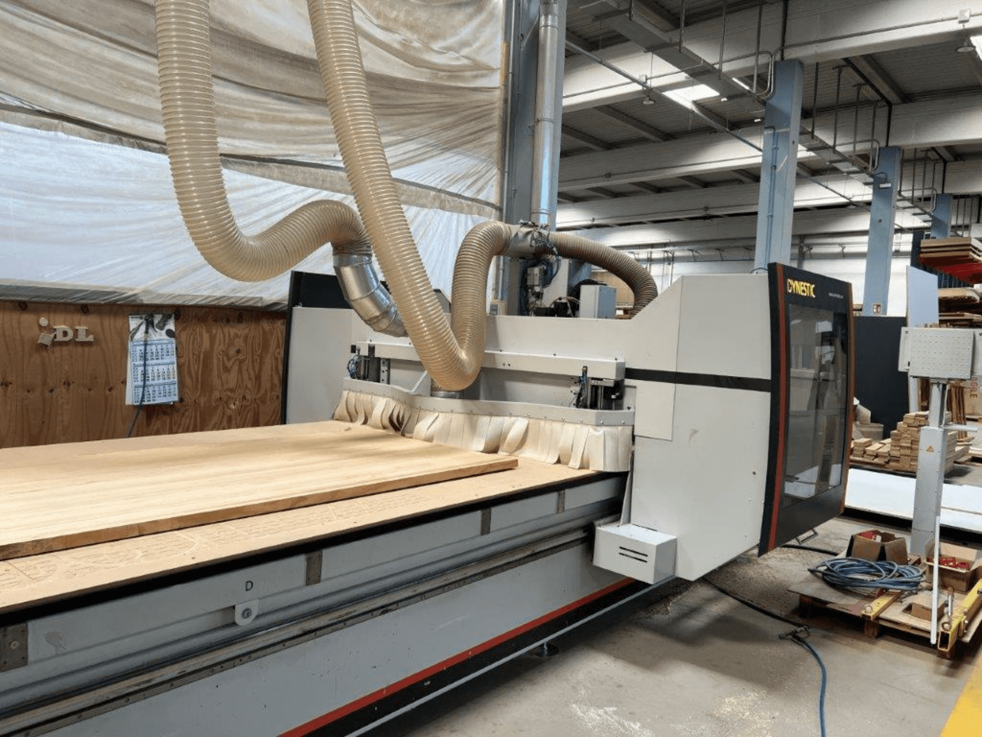 Macchina CNC di Dynestic, vista frontale, con un grande pezzo di legno e un tubo di aspirazione della polvere in un'officina.