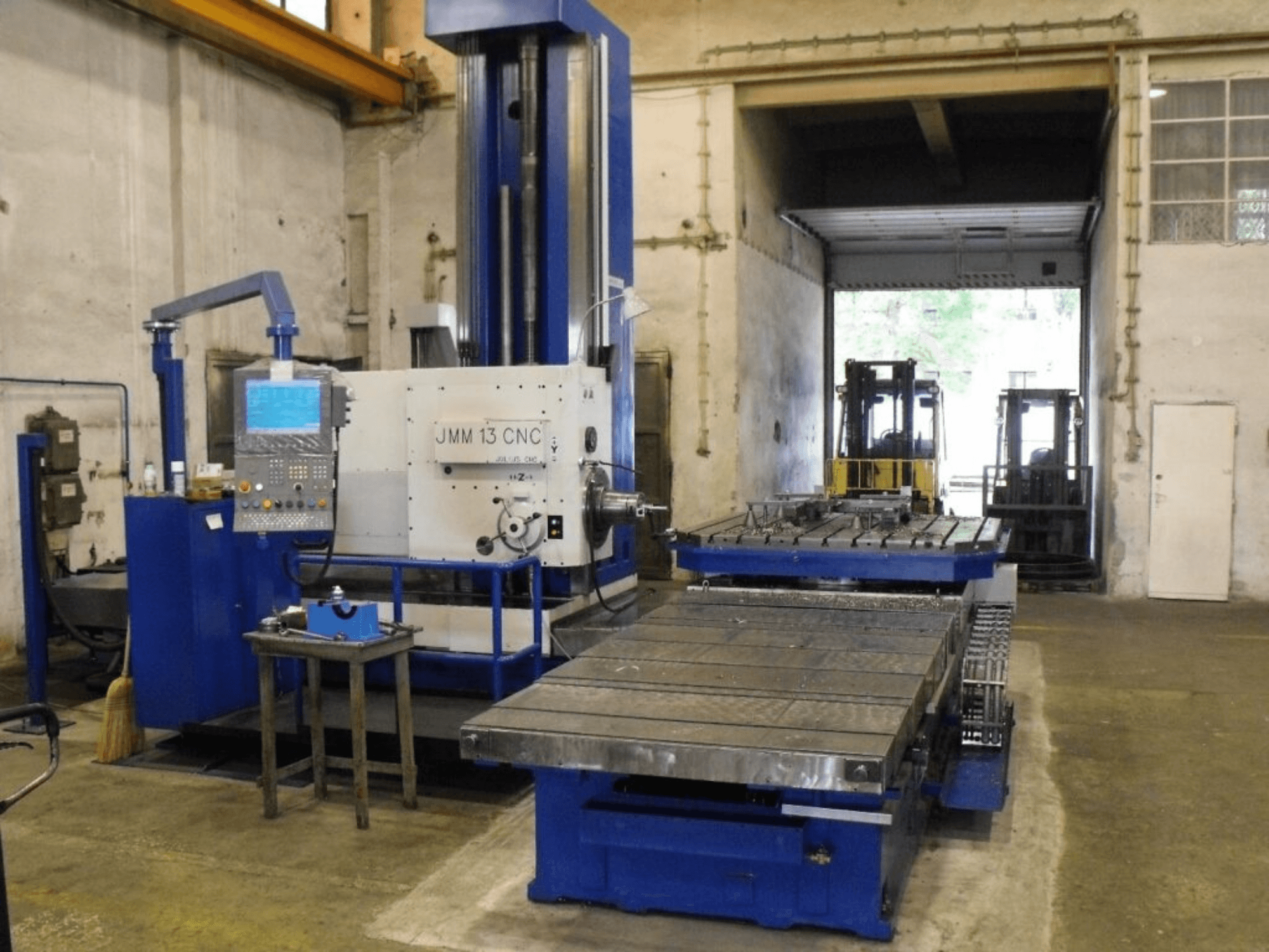 Fresatrice CNC JMM 13 in un'officina, vista frontale, con pannello di controllo, ampio tavolo di lavoro e sfondo industriale.