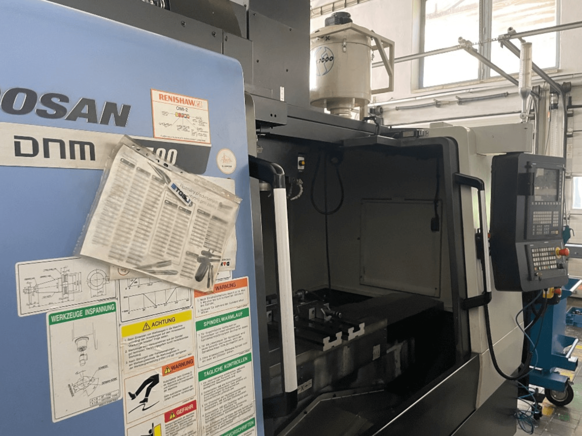 Centro di lavoro verticale DOOSAN DNM 5700, vista frontale, mostra il pannello di controllo, le etichette di sicurezza e i dettagli della macchina.