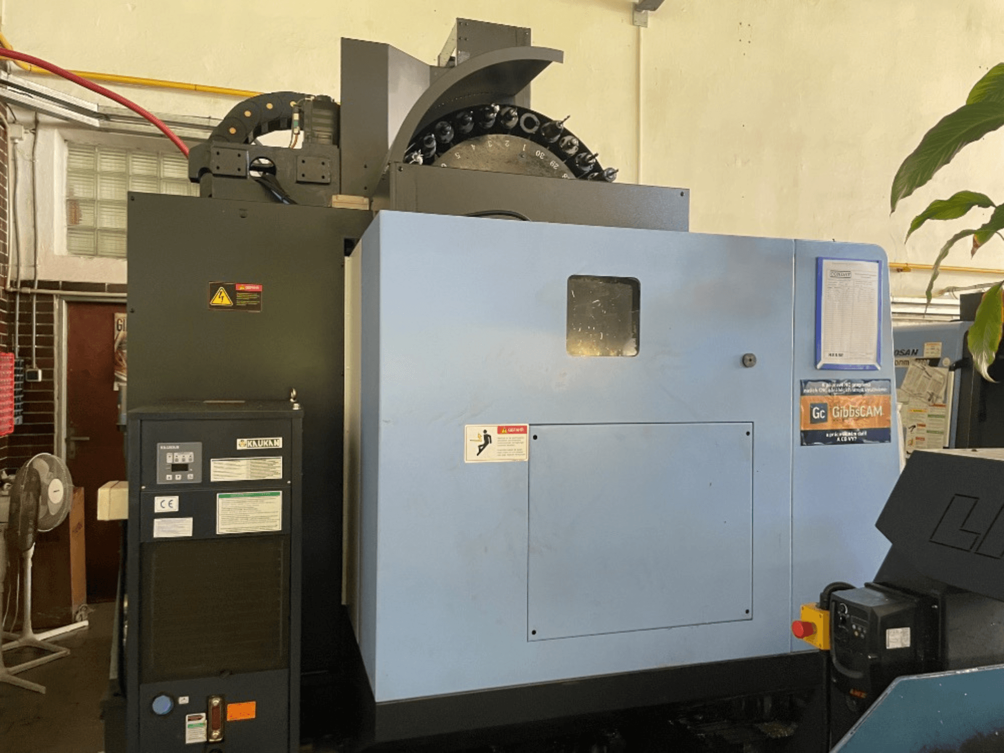 Vista frontale di un centro di lavoro verticale DOOSAN DNM 5700, con pannello di controllo, etichette di sicurezza e caratteristiche della macchina.