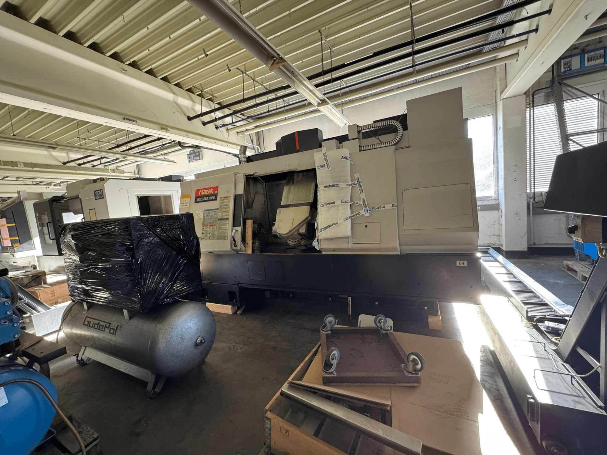 Vista frontale del centro di fresatura-tornitura Mazak INTEGREX 200-3 all’interno di un’officina, parzialmente nascosto dall’imballaggio e dalle attrezzature circostanti.