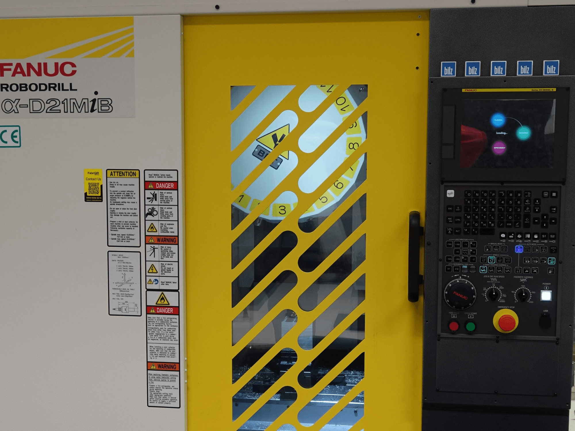 Vista frontale del centro di lavorazione verticale FANUC α-D21MiB, con le avvertenze di sicurezza, il pannello di controllo e la porta di sicurezza gialla.