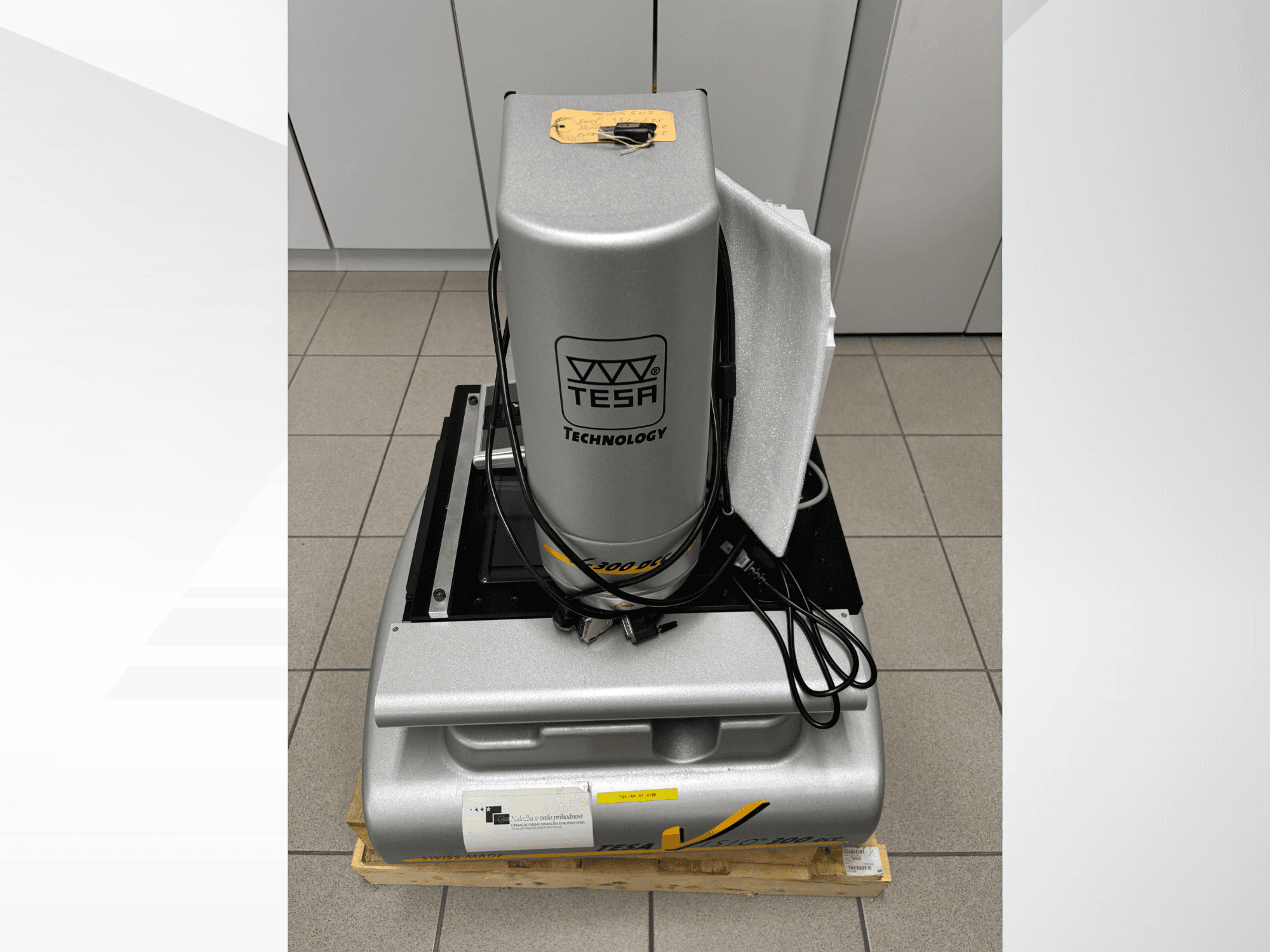 Macchina di misura ottica TESA Visio 300 DCC in vista frontale con i cavi, su un pallet di legno e con l'imballaggio protettivo visibile.