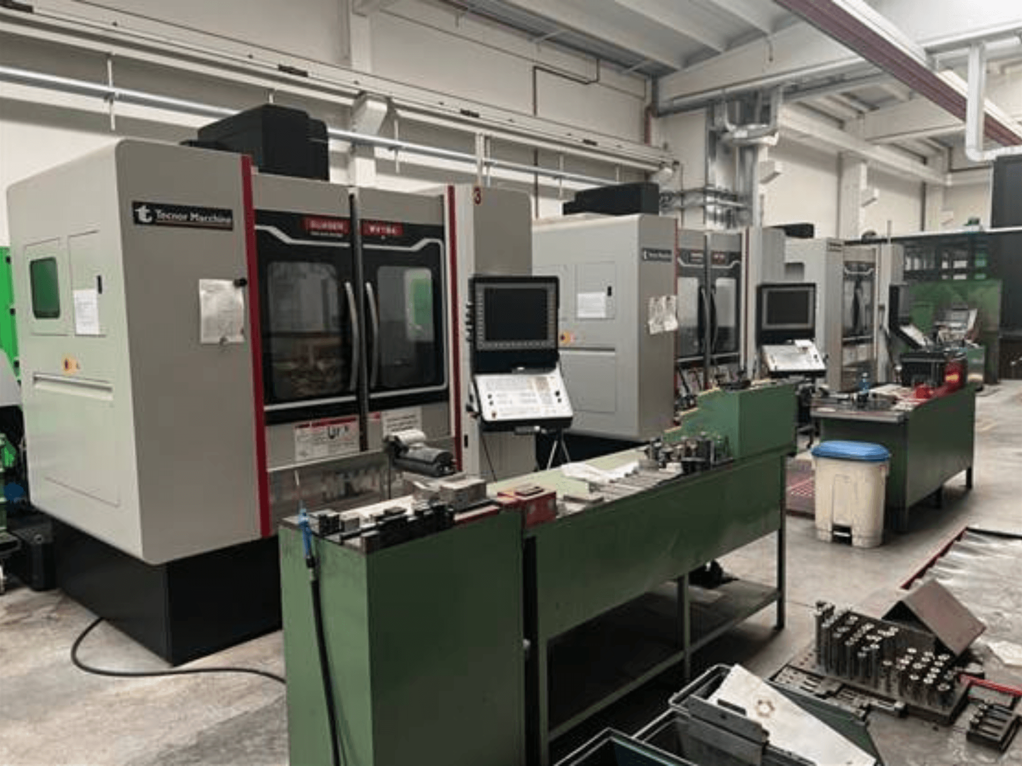 Quattro macchine CNC di Tecnoer Marchine in un'officina, vista frontale, con utensili e pezzi su banchi da lavoro verdi.