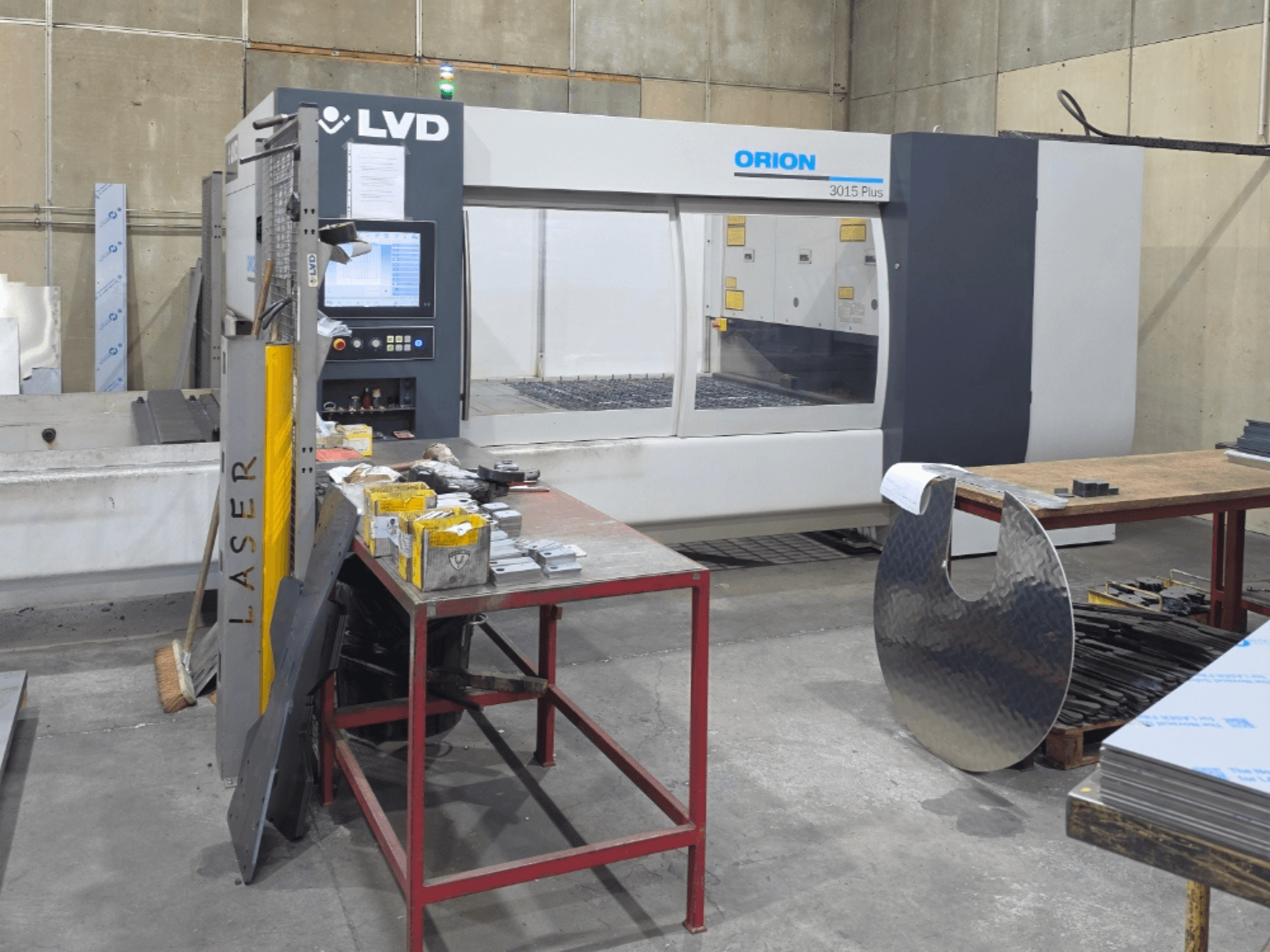 Macchina da taglio laser a CO₂ LVD Orion 3015 Plus, vista frontale con pannello di controllo; circondata da parti metalliche su un tavolo di lavoro.