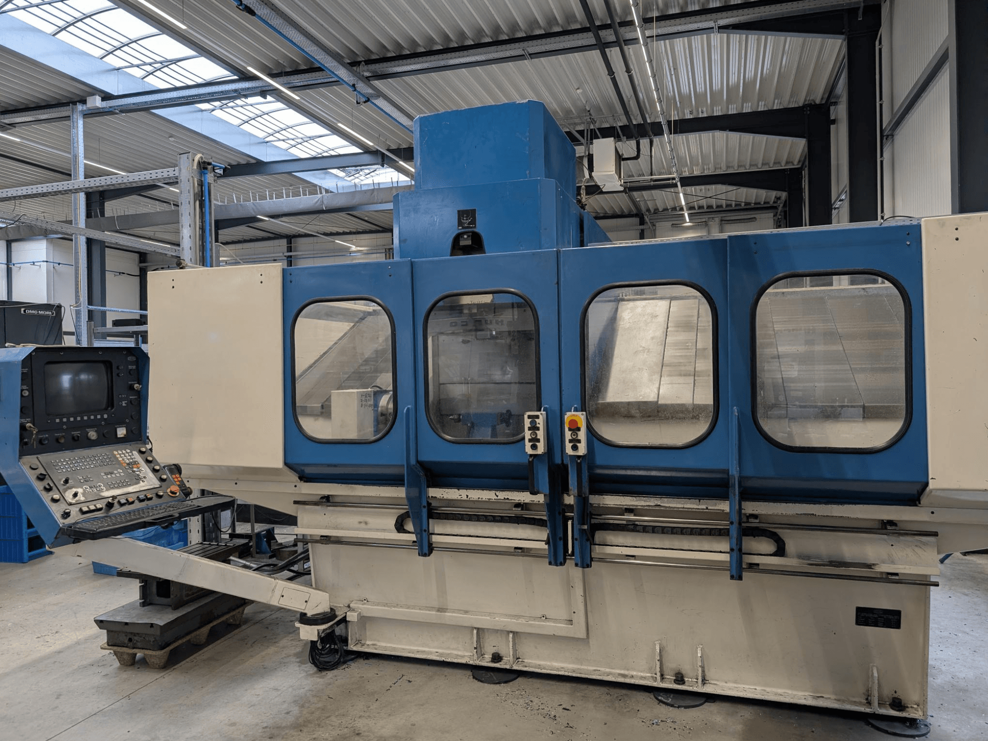 Macchina industriale CNC blu con pannello di controllo, vista frontale. Grandi finestre in vetro, componenti e pulsanti del macchinario visibili.