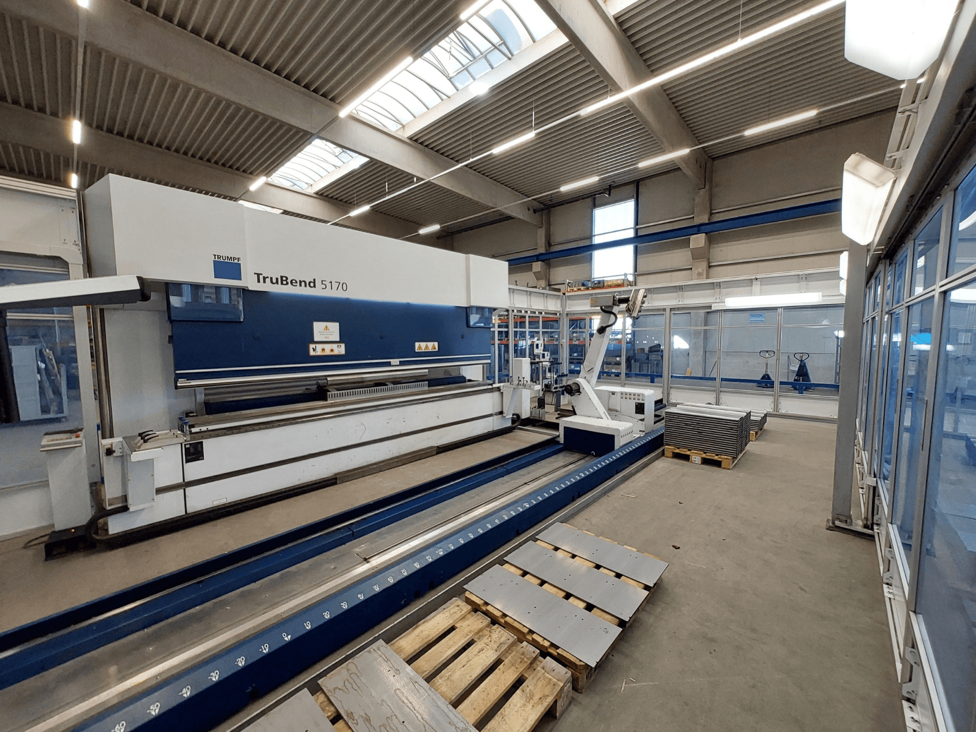 Pressa piegatrice TRUMPF TruBend 5170 in un'officina, vista laterale, che mostra l'utensile di piegatura e le lamiere impilate su pallet.