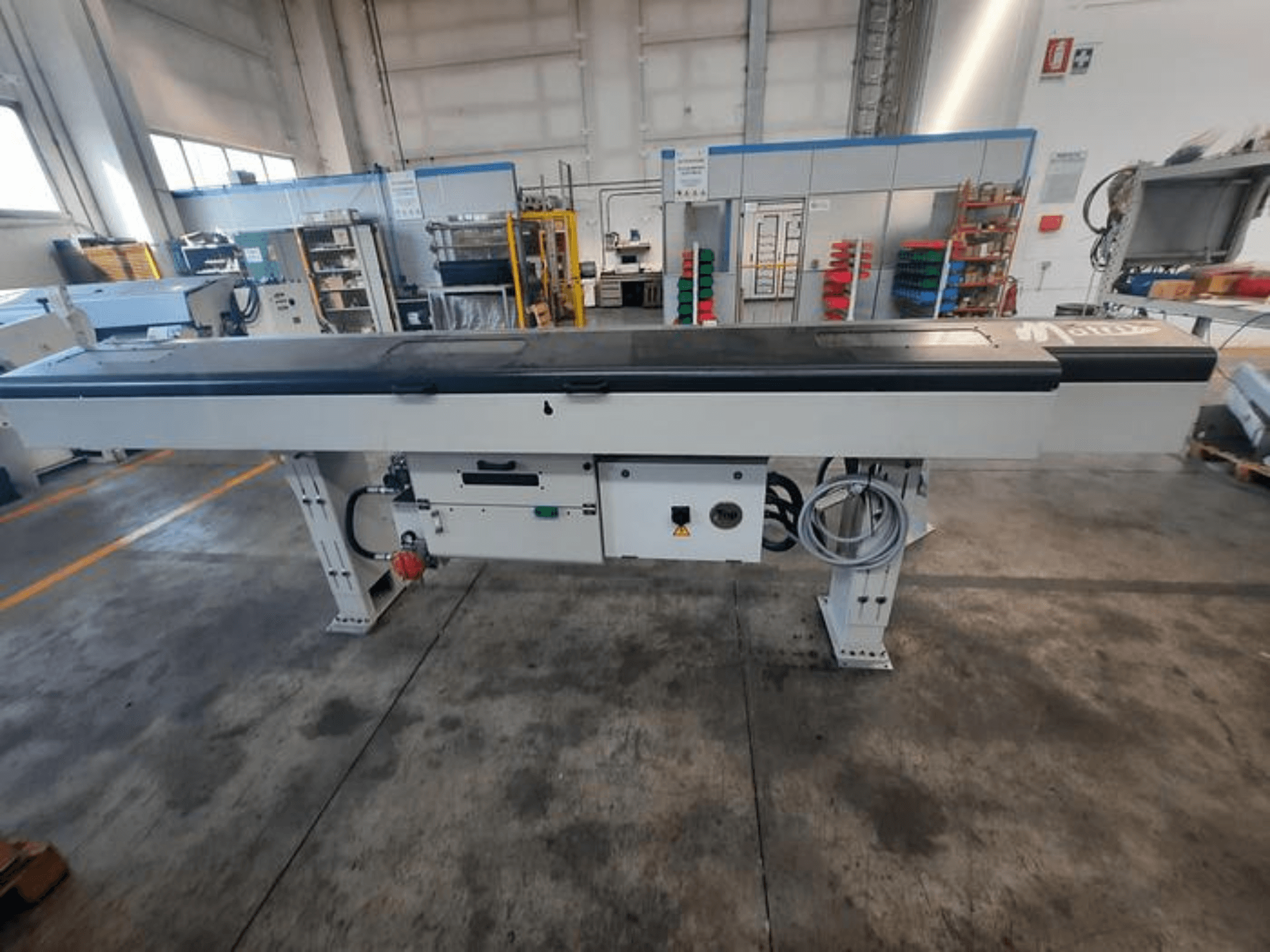 Tavolo trasportatore in metallo con piano nero liscio, vista laterale, con pulsanti di controllo e cavi; logo del marchio parzialmente visibile.