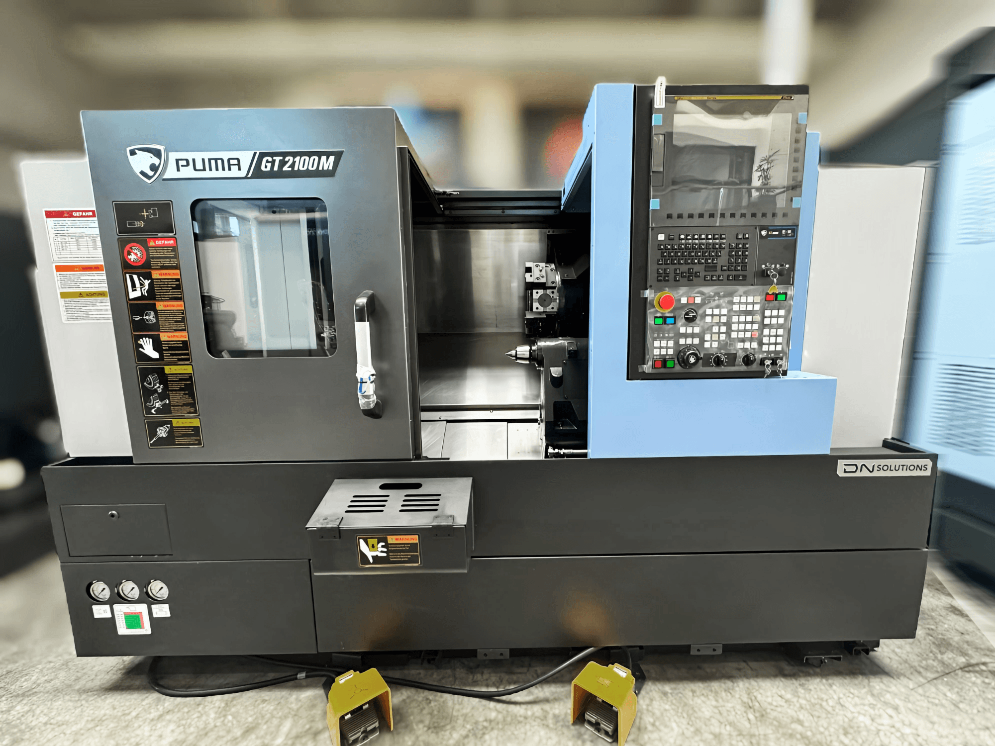 Centro di fresatura DN SOLUTIONS PUMA GT2100M, vista frontale che mostra il pannello di controllo, la camera di lavorazione e le etichette di sicurezza.