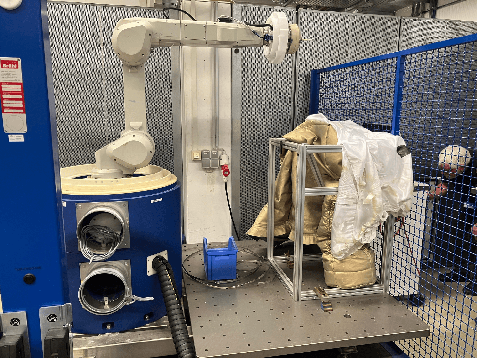 Braccio robotico industriale accanto a una macchina cilindrica blu, che tiene indumenti in tessuto su un telaio metallico, vista laterale.