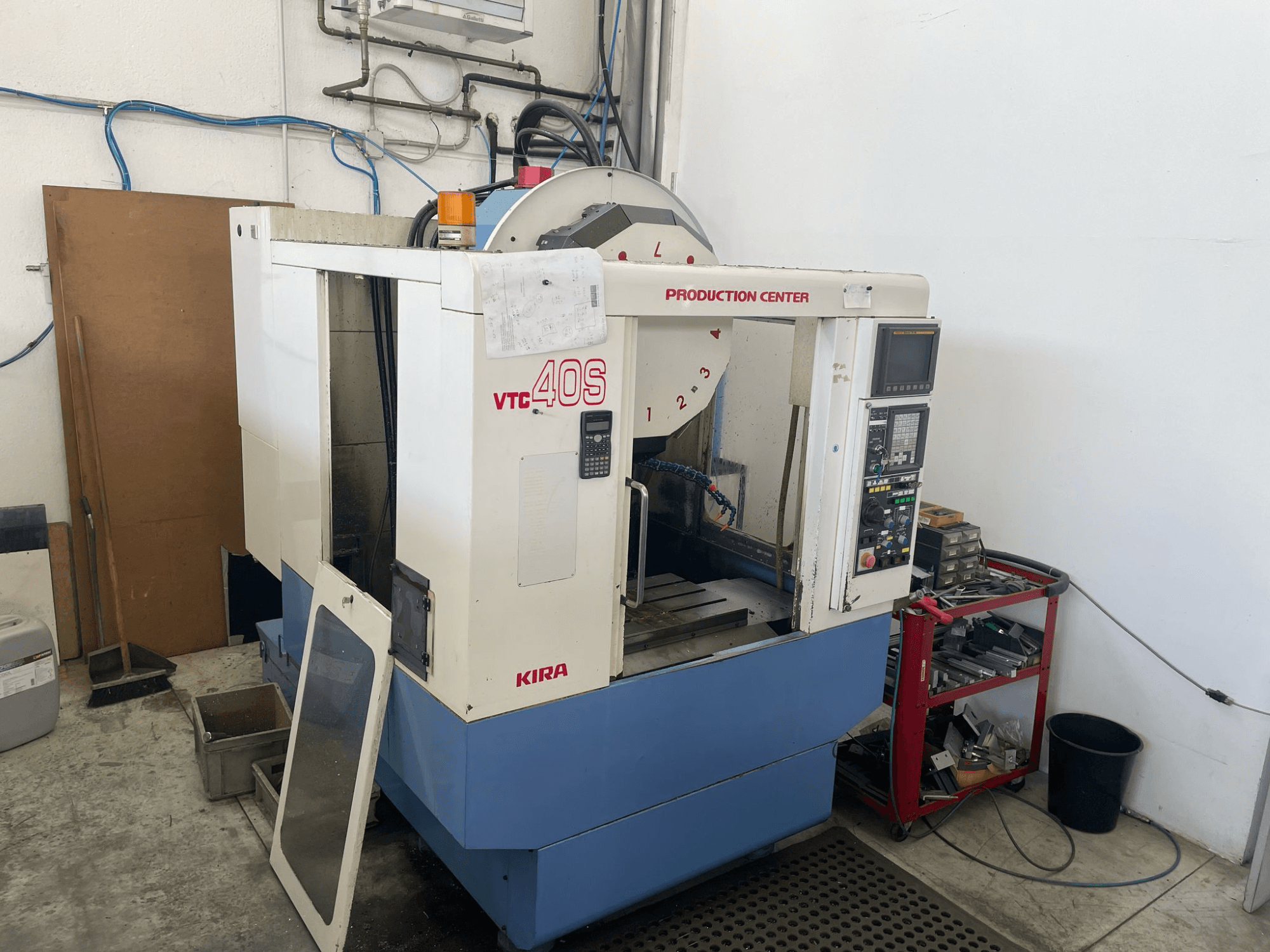 Centro di lavoro KIRA VTC 40S in vista laterale, con pannello di controllo, porta aperta e utensili su un carrello di stoccaggio vicino.