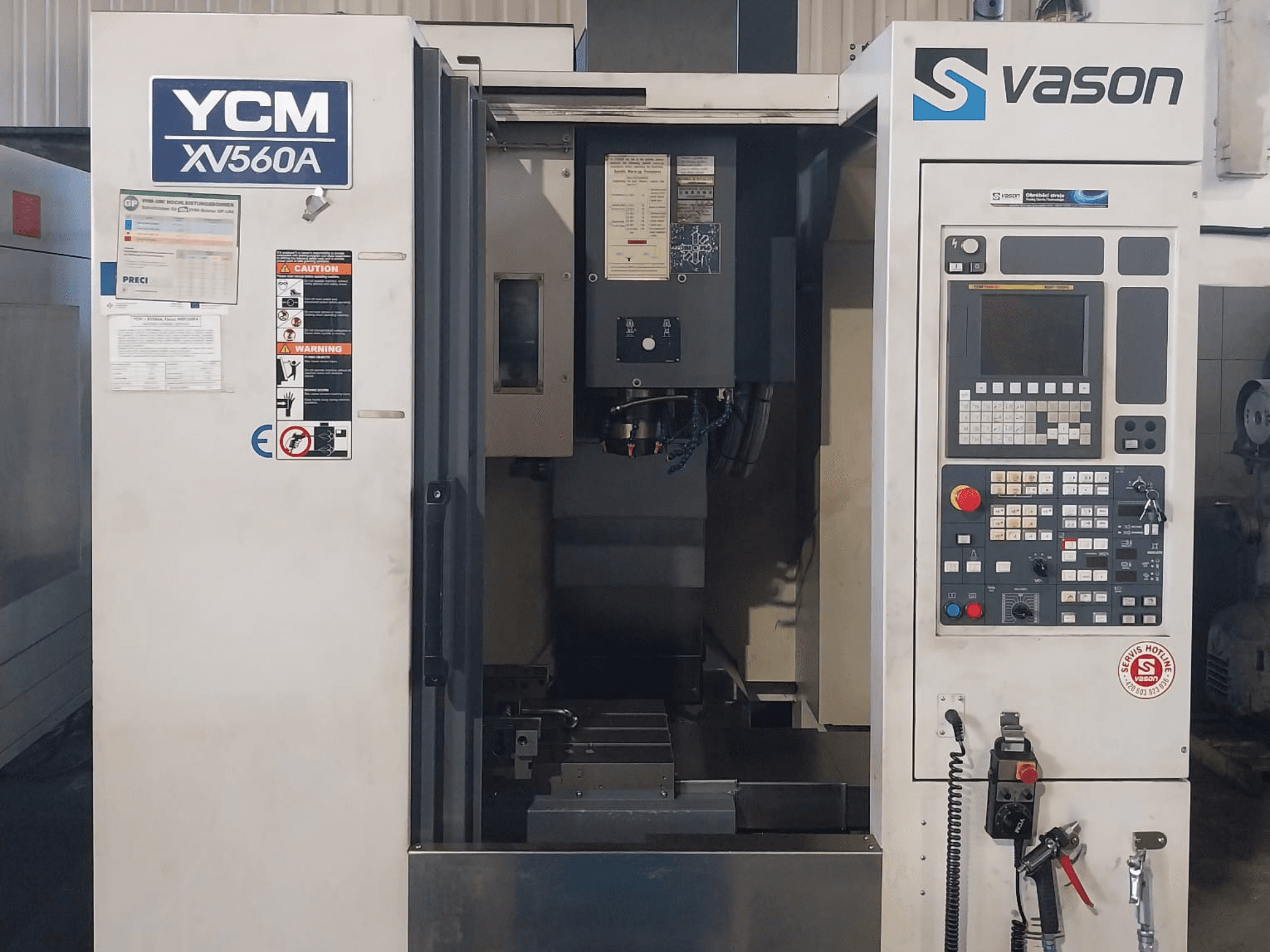 Vista frontale del Centro di lavoro verticale YCM XV 560A, con pannello di controllo e piano di lavoro con componenti di lavorazione visibili.