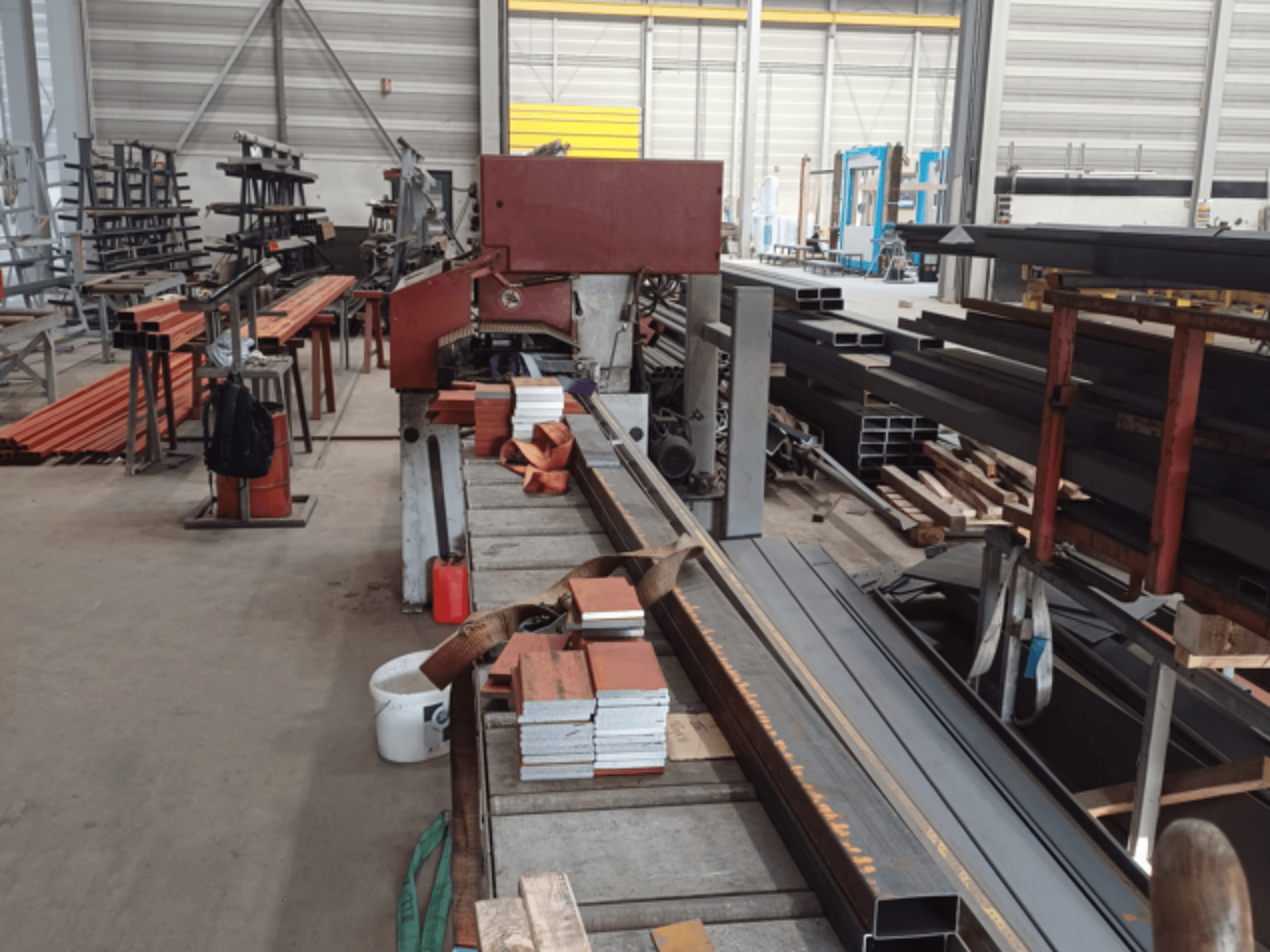 Officina di produzione di vari profili metallici, con una segatrice rossa in primo piano e materiali impilati.