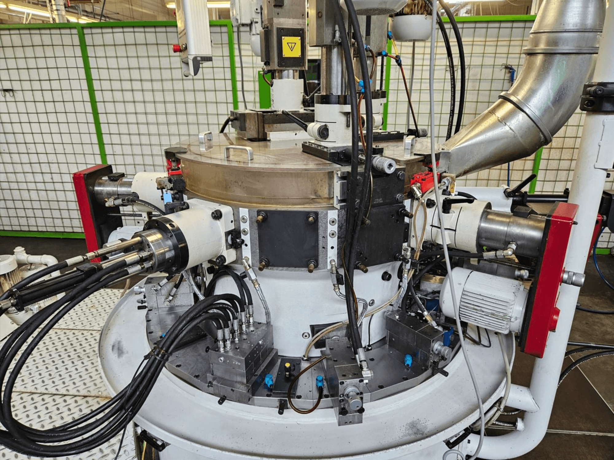 Vista ravvicinata di una macchina industriale con tubi idraulici, componenti metallici e una base rotante. Non è visibile il marchio.