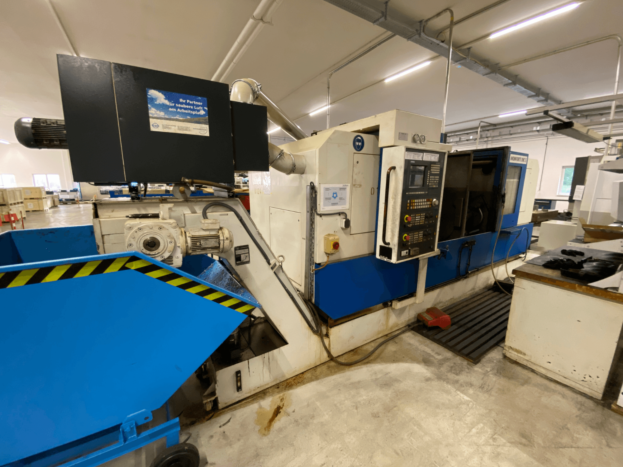 Centro di tornitura e fresatura Monforts DNC 5, vista laterale che mostra il pannello di controllo, il vassoio per i trucioli blu e i dettagli del macchinario in un ambiente di officina.
