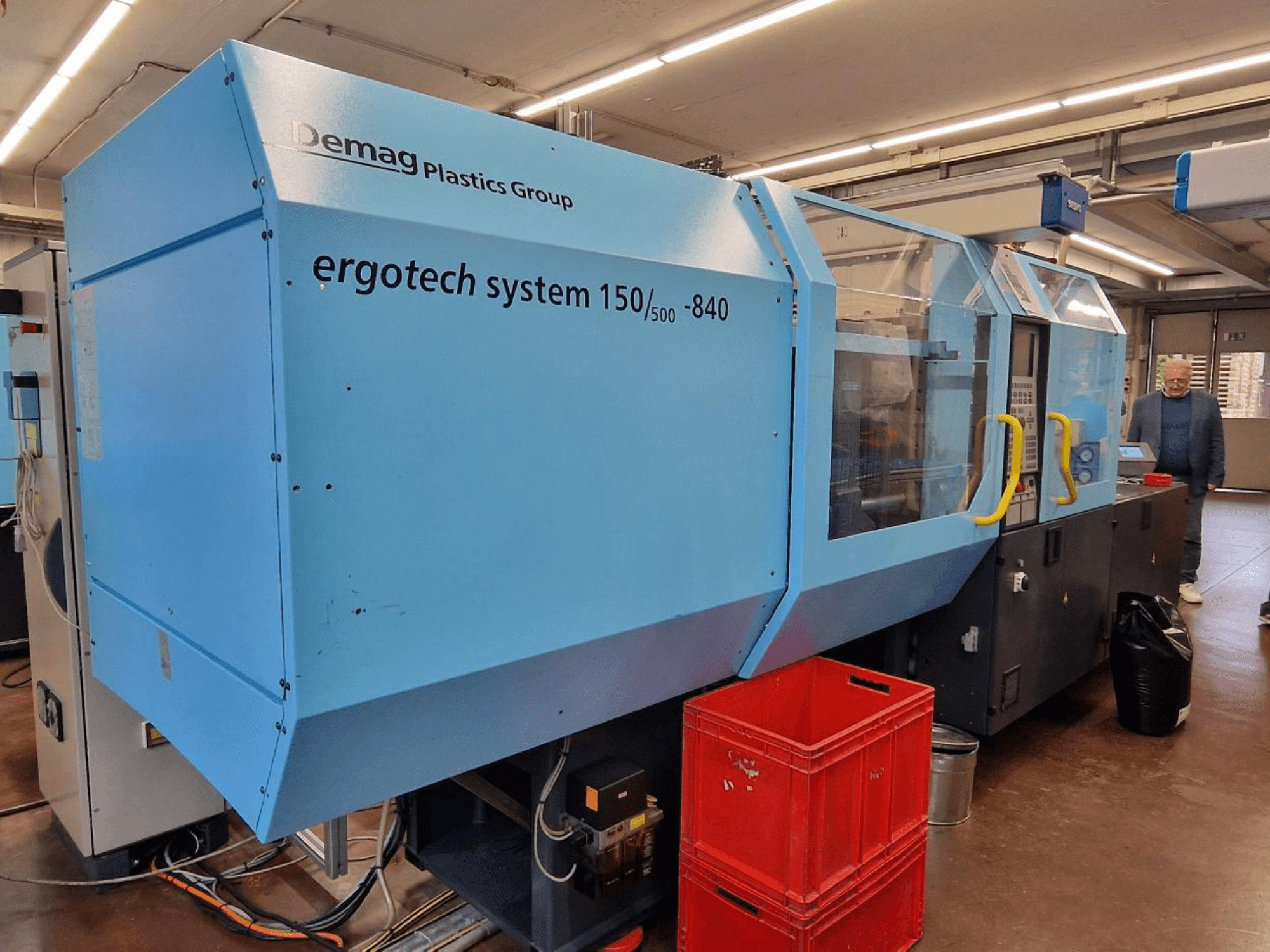 Macchina ad iniezione Demag Ergotech System 150/500-840 in blu, vista laterale, con casse e operatore sullo sfondo.