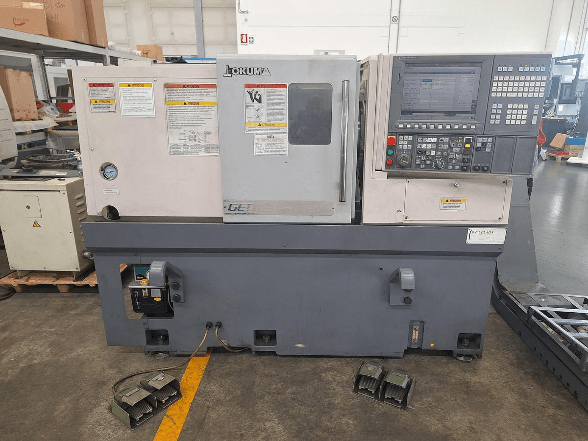 Tornio orizzontale Okuma GENOS L200E-M in vista laterale; pannello di controllo, avvisi di sicurezza e pedali.