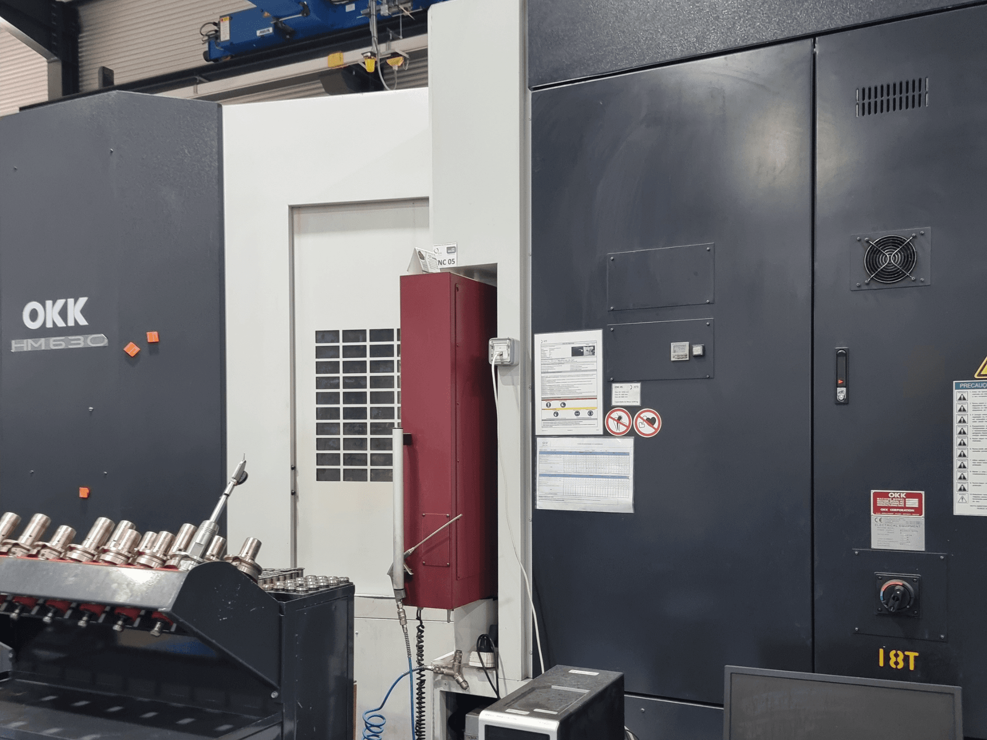 Centro di lavorazione orizzontale OKK HM630, vista frontale che mostra il pannello di controllo, le avvertenze di sicurezza e il vano porta-utensili sul lato.