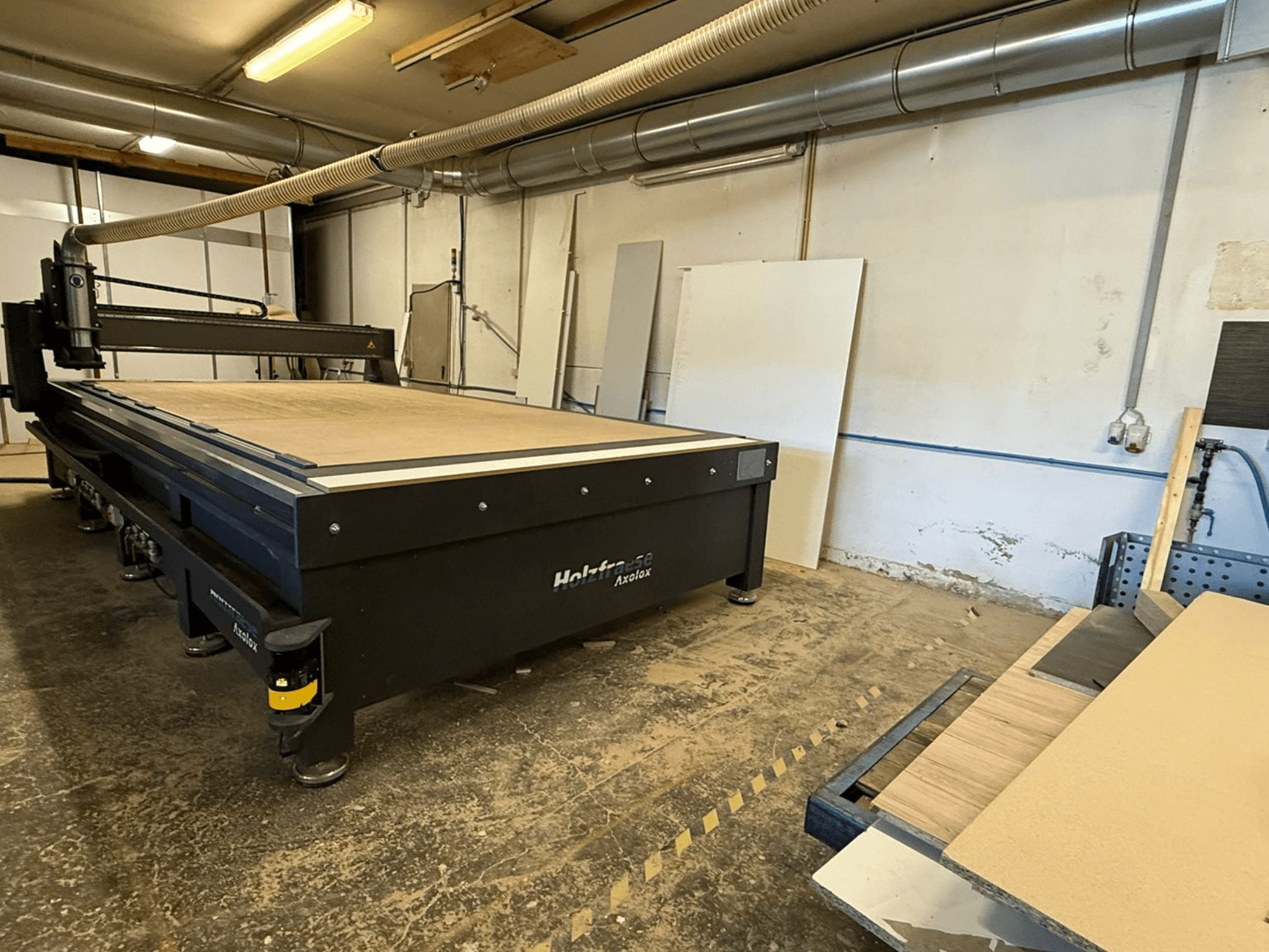 Pantografo CNC Holz-Her Axos 5 in officina, vista frontale che mostra il piano di lavoro, il sistema di aspirazione della polvere e i pannelli di legno nelle vicinanze.