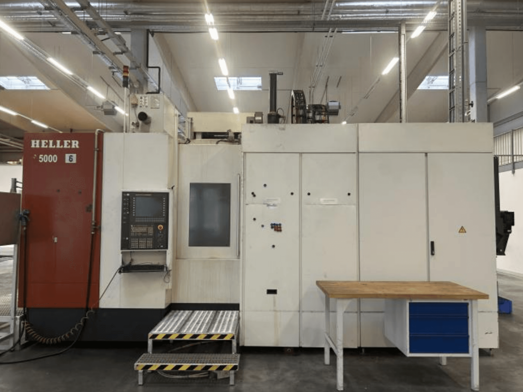 Vista frontale del centro di lavorazione orizzontale HELLER H5000 con pannello di comando, piattaforma rialzata e tavolo di lavoro adiacente.