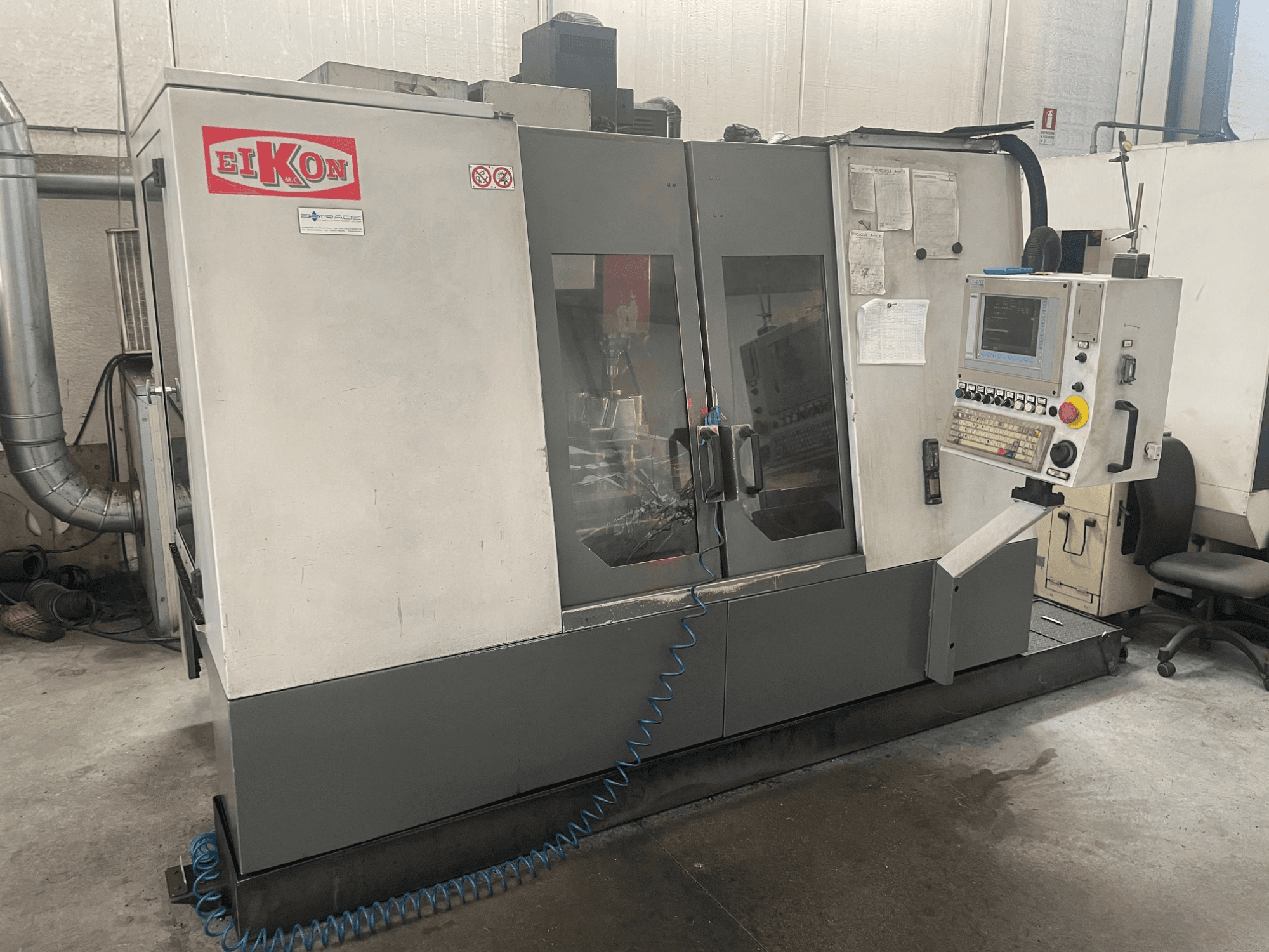 Macchina CNC EIKON vista frontalmente, con pannello di controllo, grandi porte in vetro e tubo dell'aria blu sul pavimento.