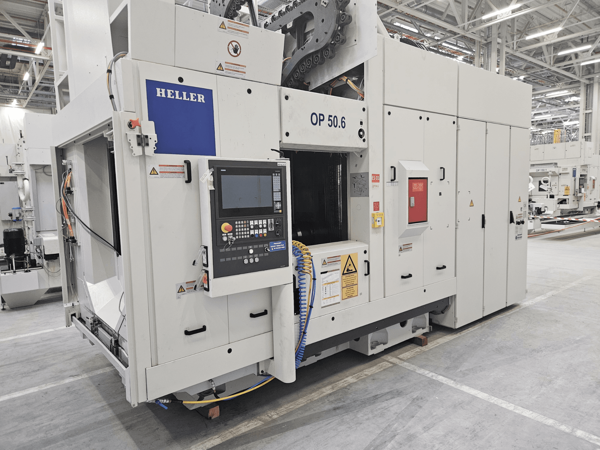 Centro di lavoro Heller OP 50.6, vista frontale, con pannello di controllo, etichette di avvertenza e tubi flessibili visibili in un ambiente di fabbrica.