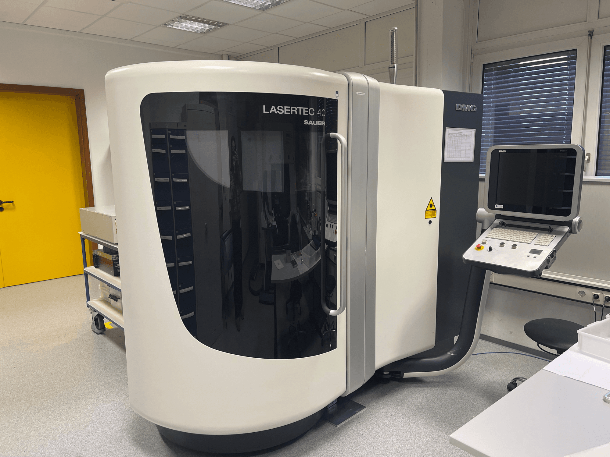 Fresatrice LASERTEC 40 bianca di DMG MORI, vista frontale, con pannello di controllo e design ergonomico in una stanza ben illuminata.