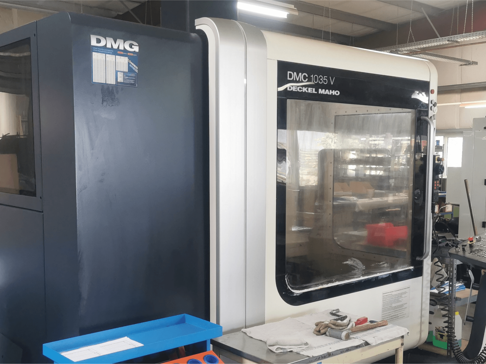 Fresatrice DMG DMC 1035 V in un'officina, vista frontale con vassoio portautensili blu e guanti sulla superficie.