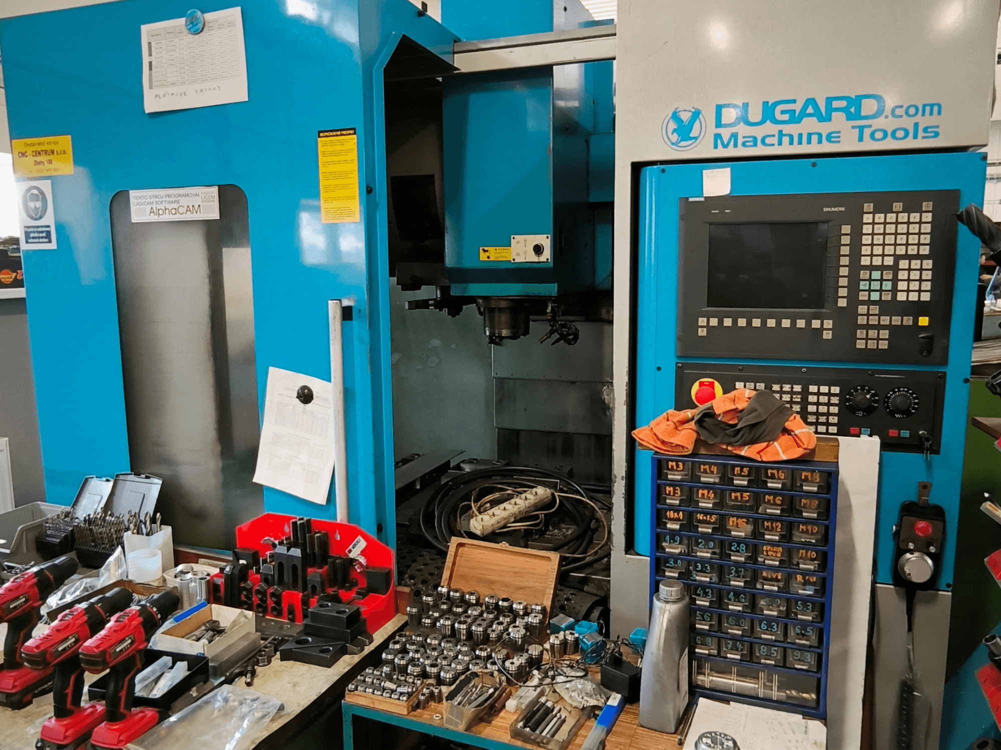 Vista frontale del centro di lavoro verticale Dugard 850VMC, con pannello di controllo, utensili e accessori su un banco di lavoro.
