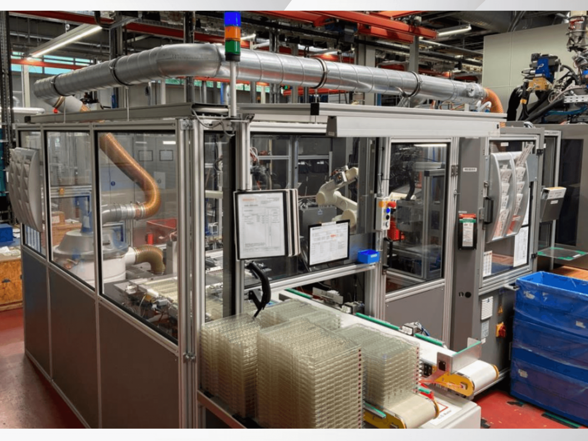 Macchina di assemblaggio automatizzata con braccio robotico, pannelli laterali trasparenti e contenitori impilati di articoli trasparenti, vista frontale.