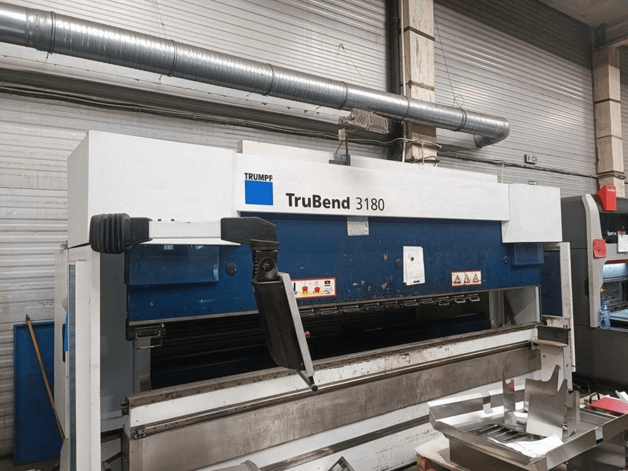 Curvatrice Trumpf TruBend 3180 in un'officina, vista laterale che mostra il pannello di controllo e i pezzi di metallo sul tavolo.