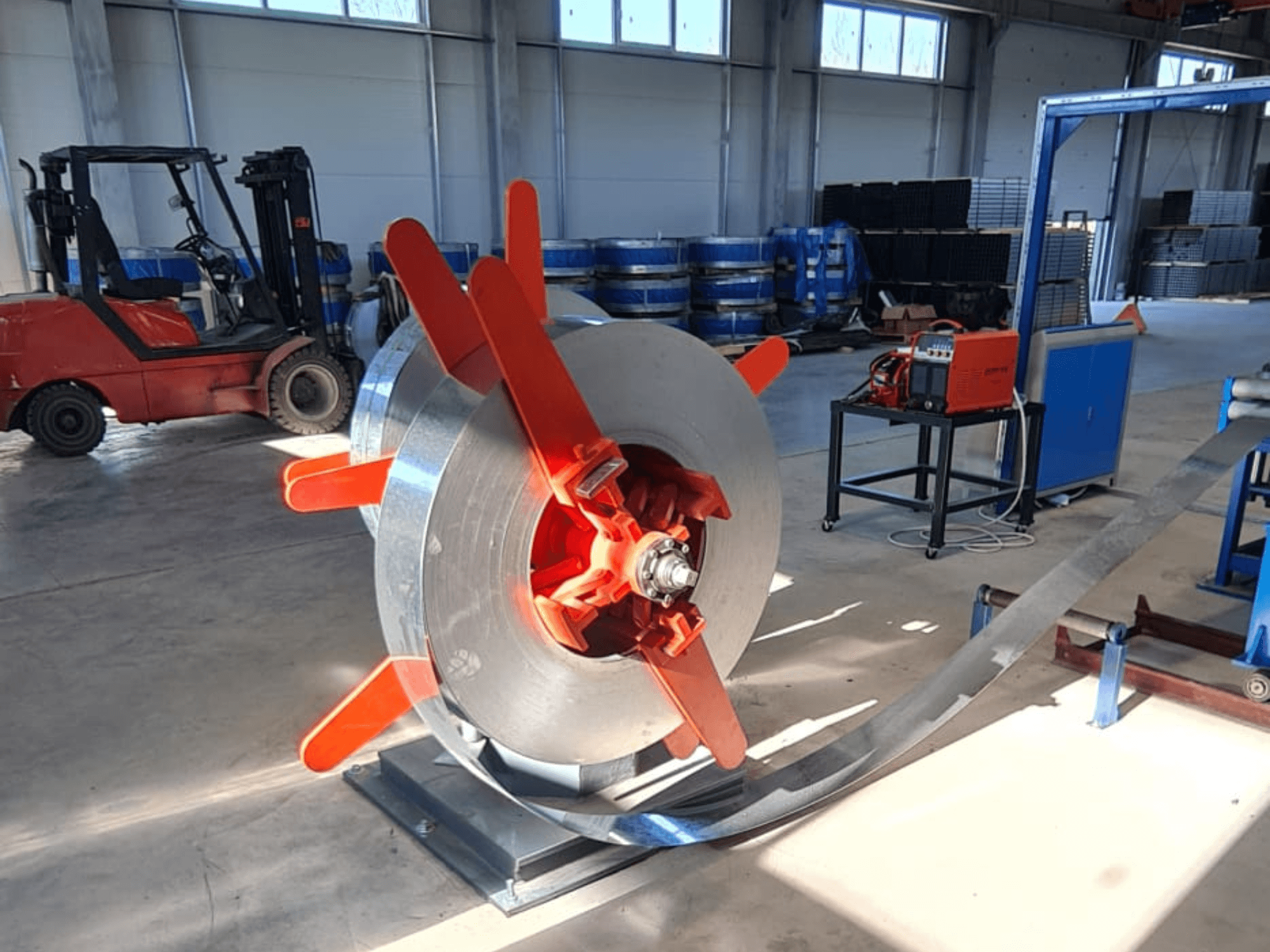 Bobina di metallo con maniglie rosse su un supporto, vista da un'angolazione. Sfondo industriale con macchinari e carrello elevatore.