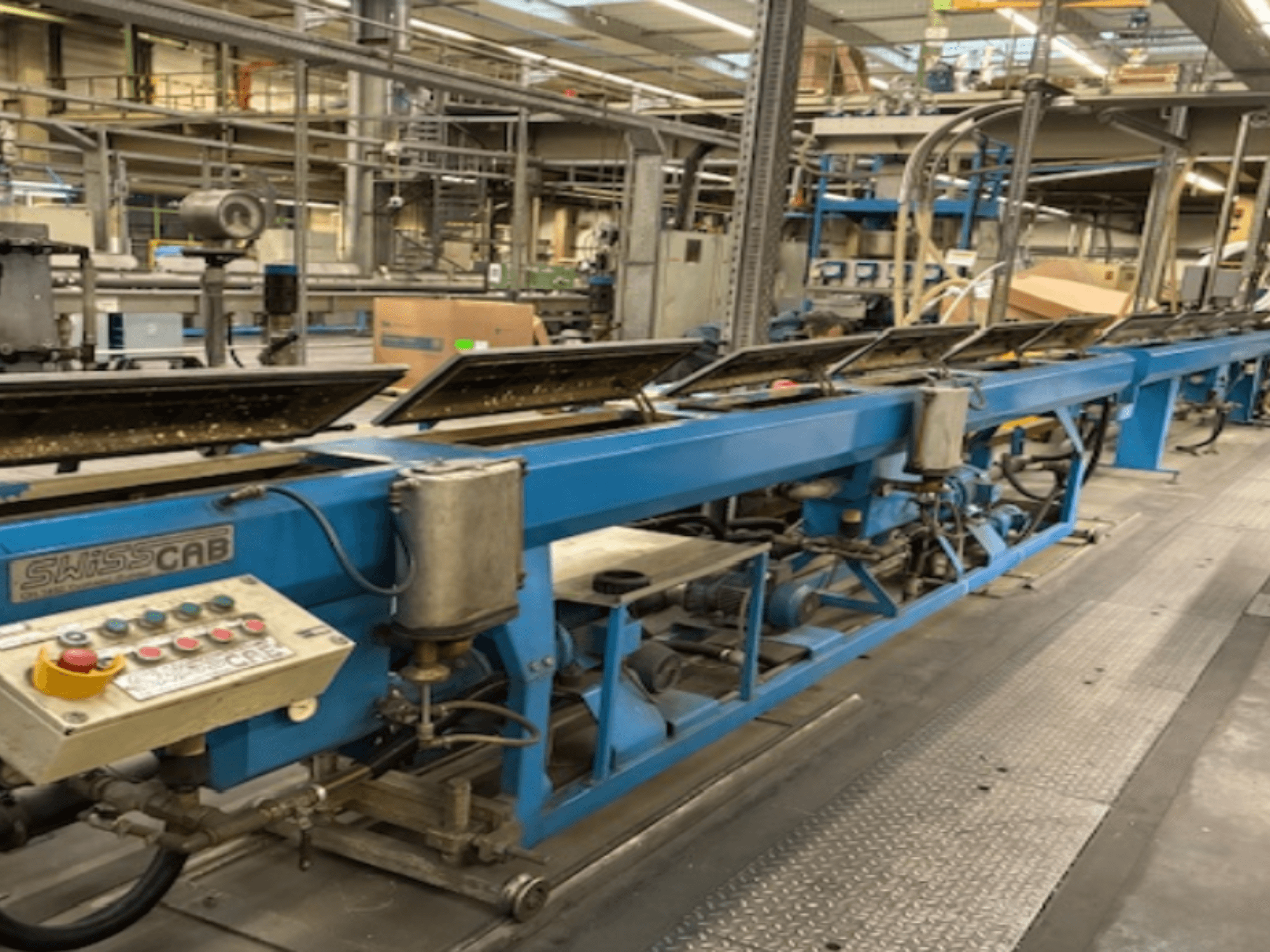 Macchina industriale SwissCab blu in un ambiente di fabbrica, con pulsanti operativi e sistema di trasporto, vista laterale.