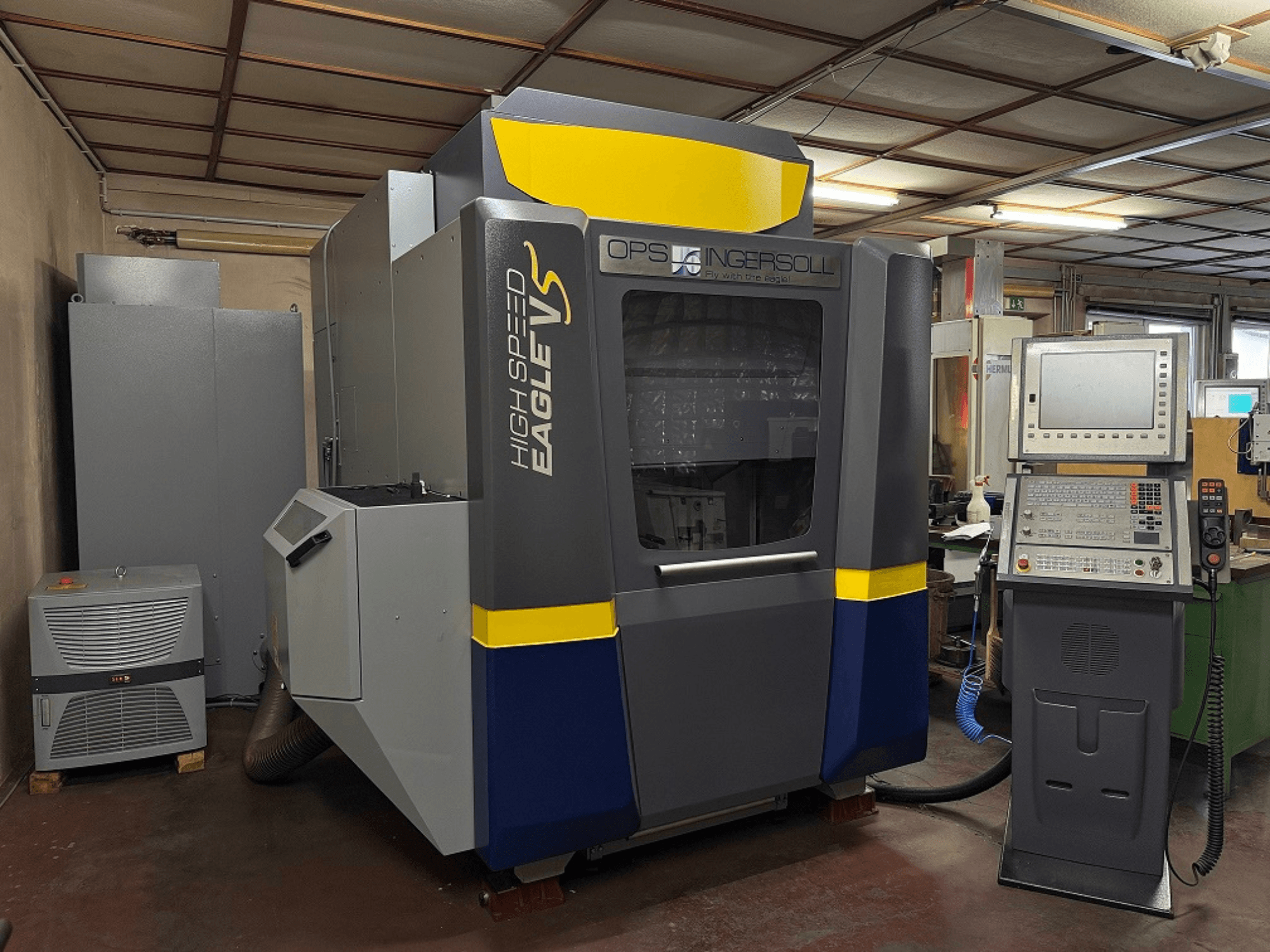 Macchina ad alta velocità Eagle V di OPS Ingersoll, vista laterale, con pannello di controllo e attrezzature adiacenti in un ambiente industriale.