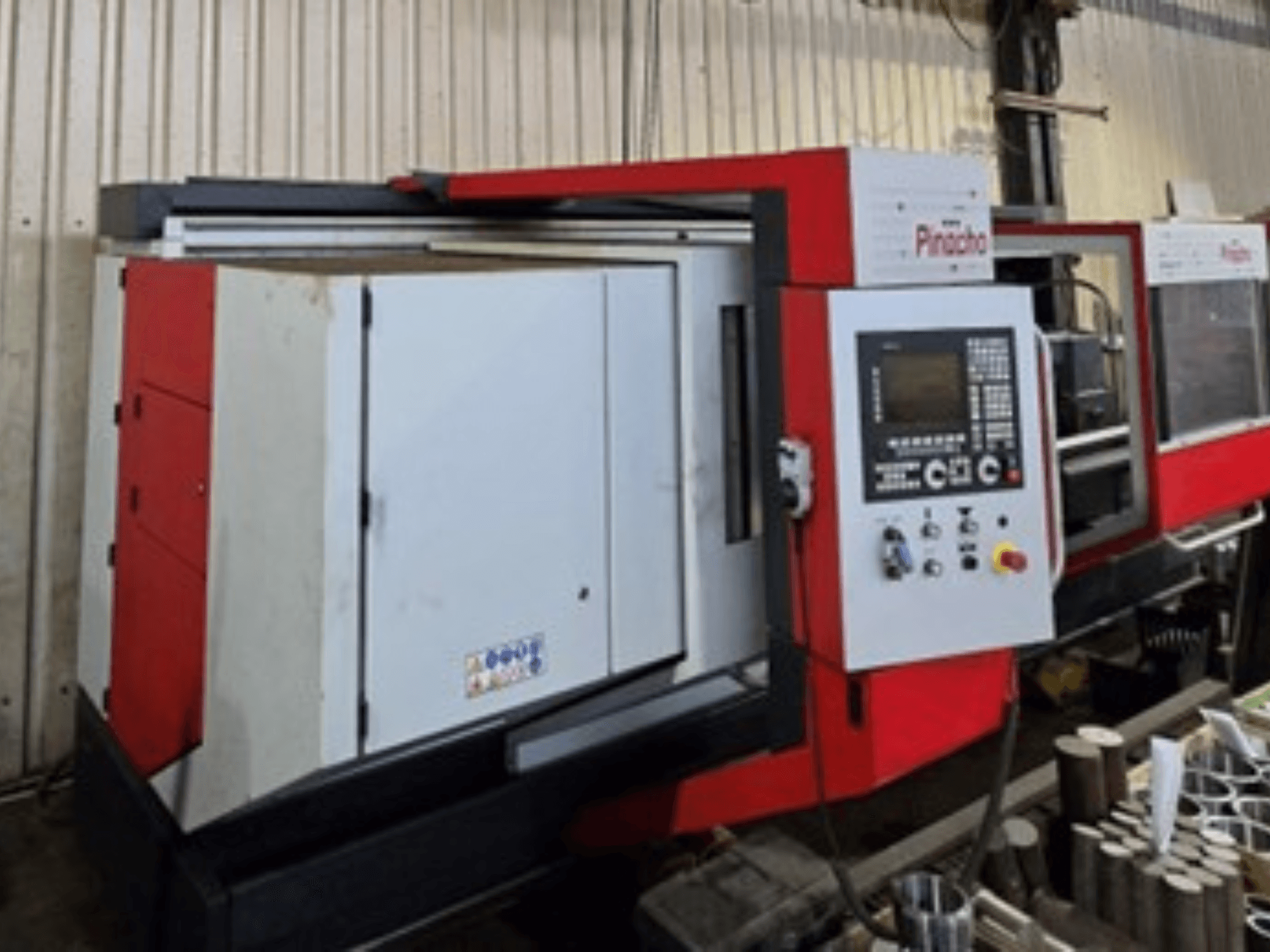 Macchina CNC Pinacho in rosso e bianco, vista laterale, con pannello di controllo e materiali sul pavimento, che mostra la configurazione operativa.