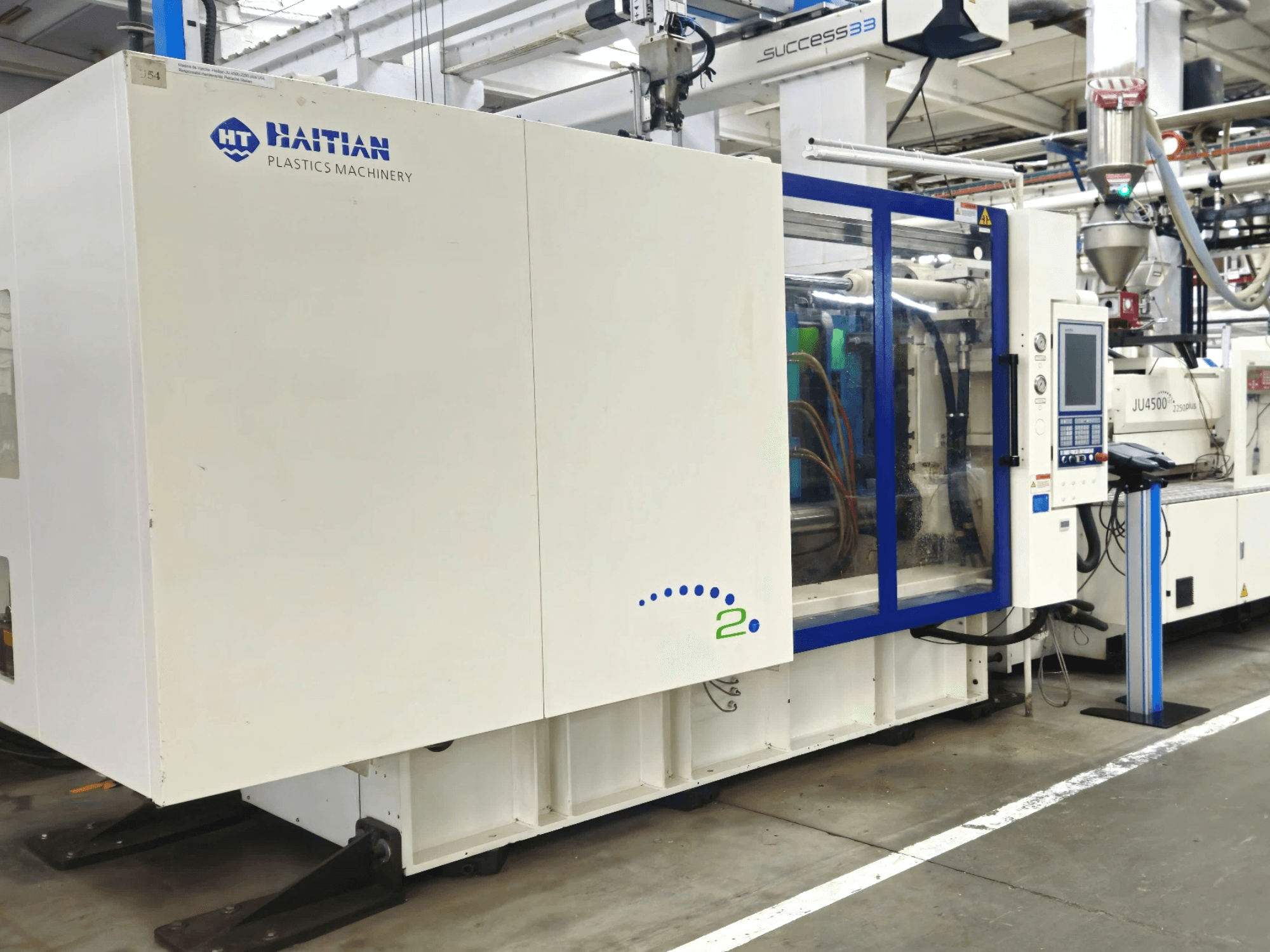 Modello di Haitian Plastics Machinery, vista frontale, che mostra un macchinario con pannello di controllo e tubi a vista, in un ambiente industriale.