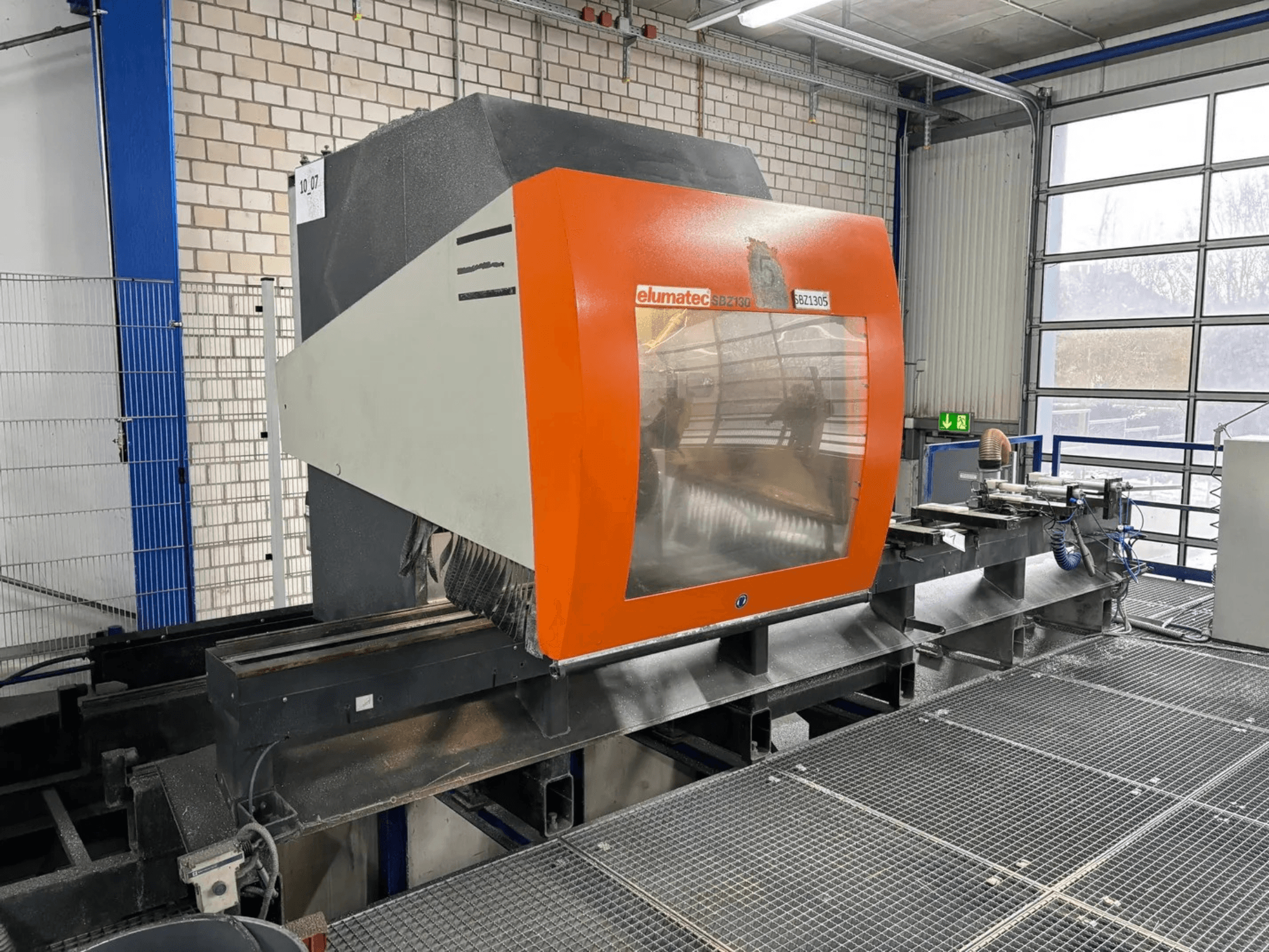 Centro di lavoro verticale Elumatec SBZ 130, vista frontale, caratterizzato da un alloggiamento arancione e grigio con un pannello trasparente.