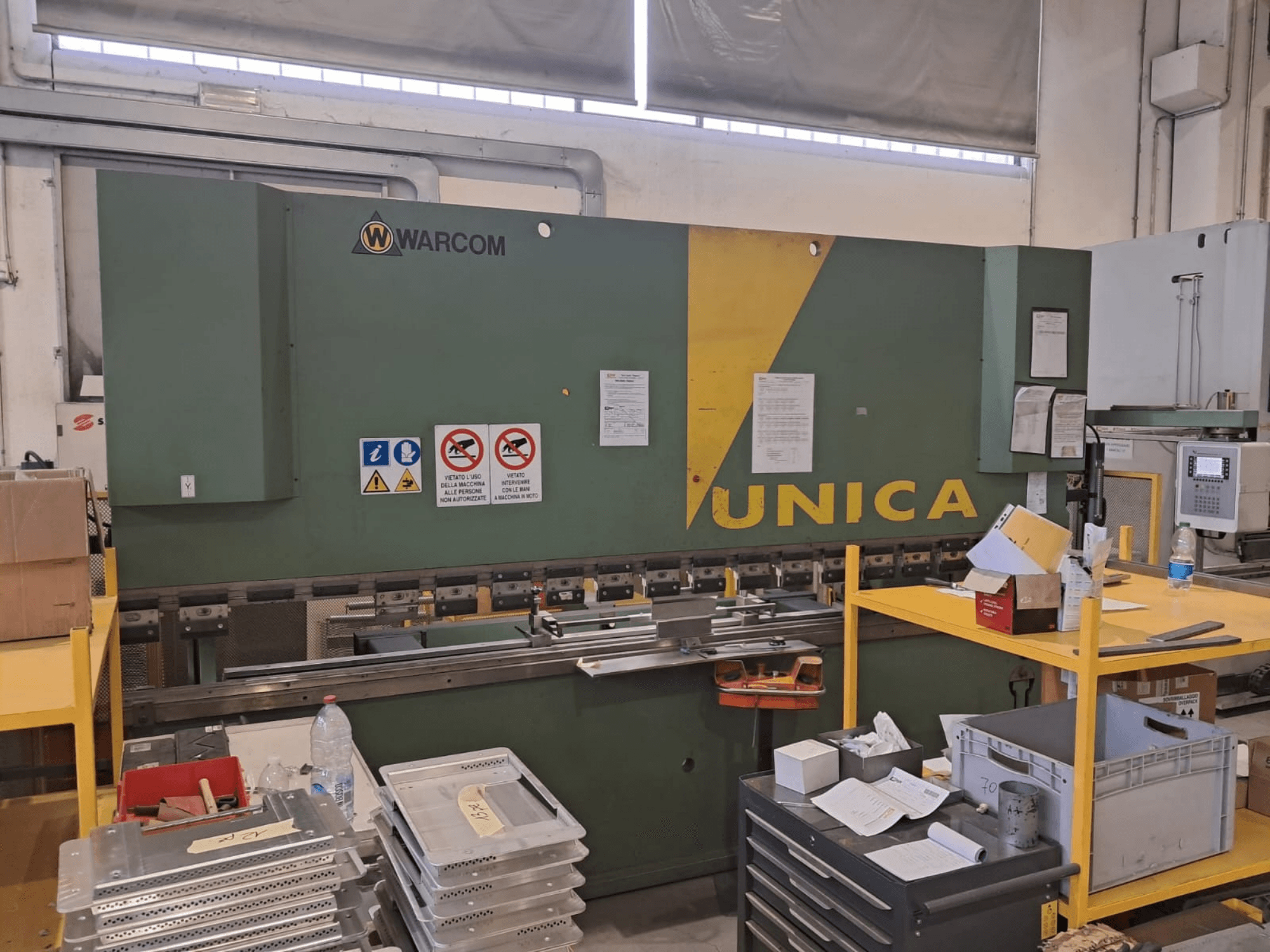 Pressa idraulica Warcom UNICA 100-40, vista frontale, corpo verde con dettagli gialli, segnaletica di sicurezza visibile, utensili e materiali sul tavolo.