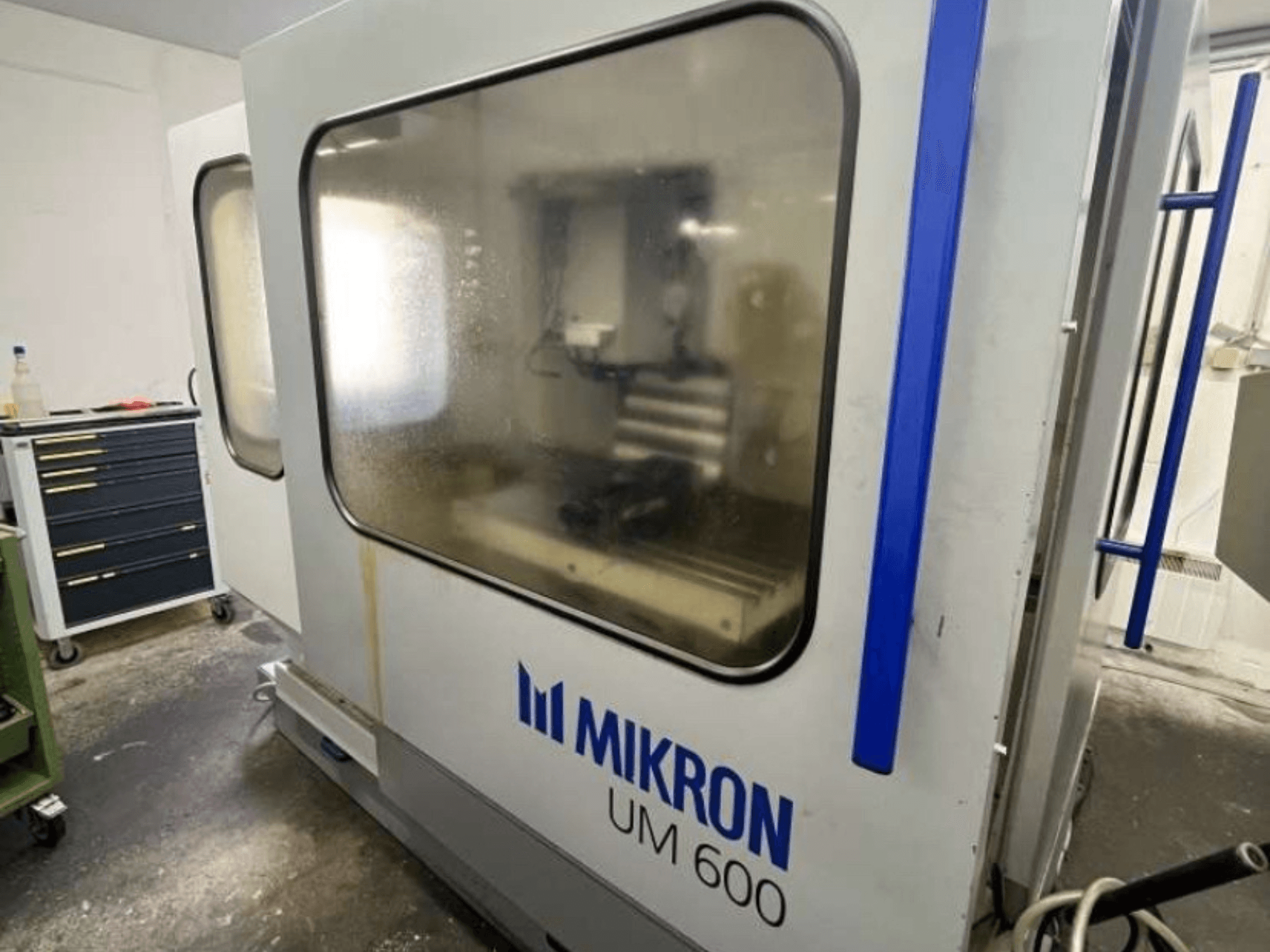 Centro di lavoro universale MIKRON UM 600 TNC 410/426, vista laterale, con vetro appannato e magazzino utensili visibile nelle vicinanze.