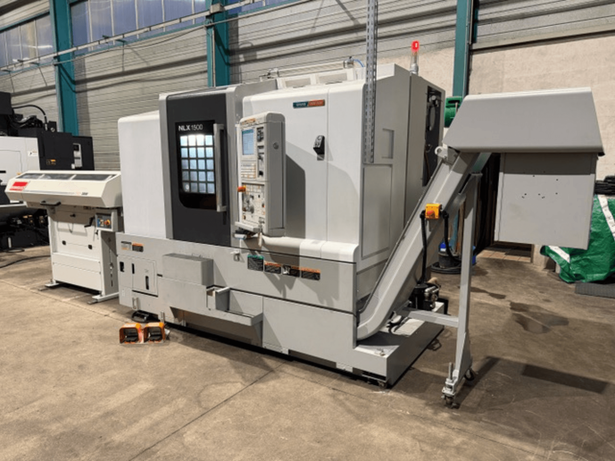 Macchina CNC NLK 1500, vista frontale, con pannello di controllo e tramoggia accessori sul lato, appoggiata su pavimento in cemento.