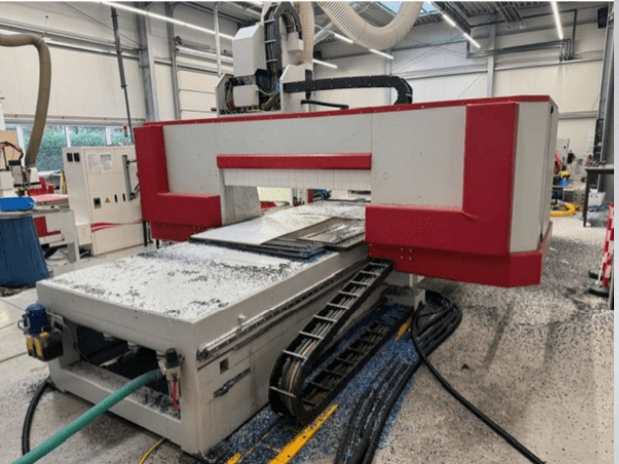 Macchina CNC rossa e grigia in un'officina, vista laterale che mostra il tavolo del materiale, il tubo del refrigerante e la segatura sul pavimento.