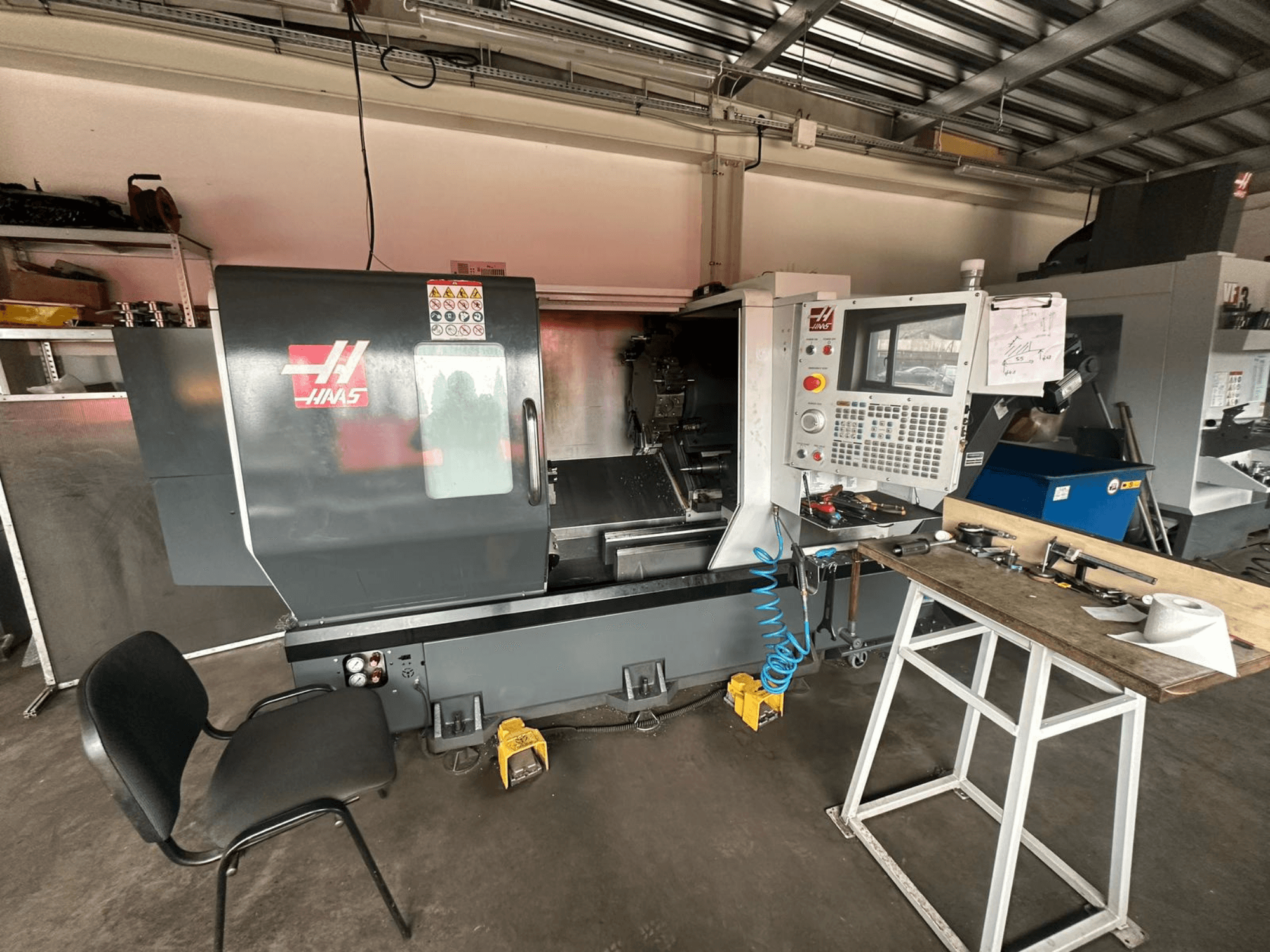 Tornio CNC Haas (modello ST-10) in un'officina, vista laterale che mostra i comandi, gli utensili su un banco di lavoro e una sedia nelle vicinanze.